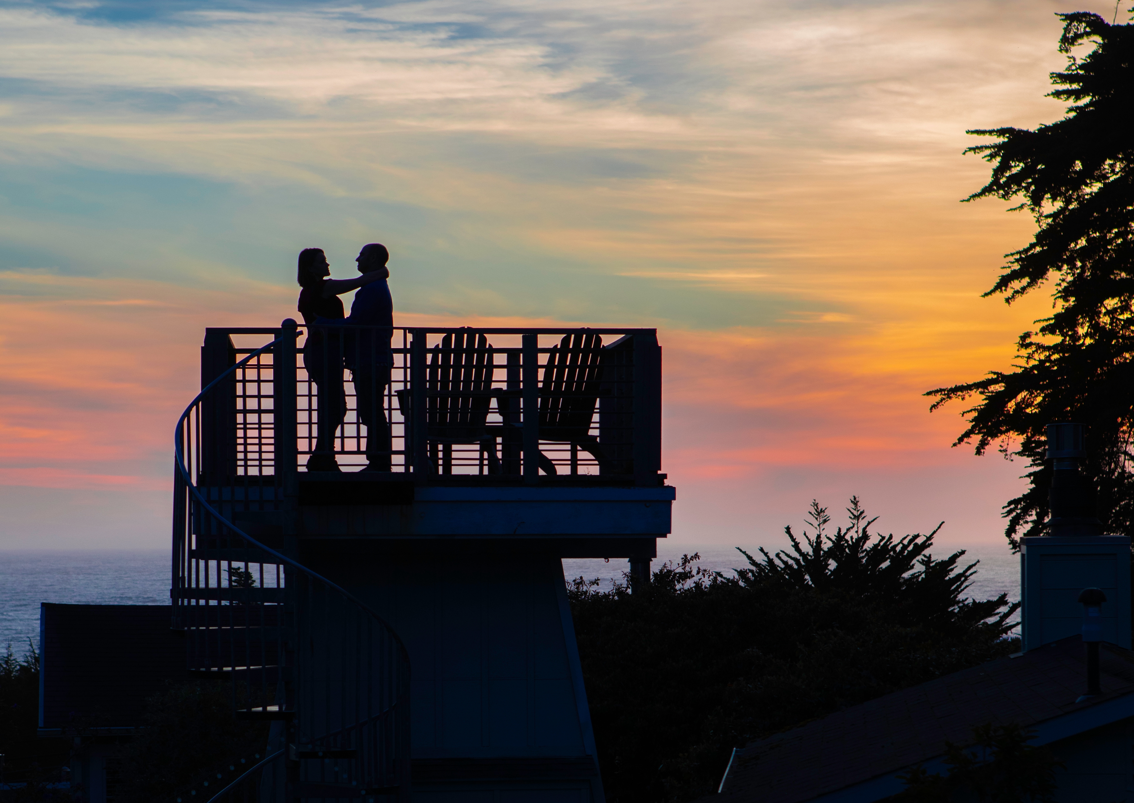 Two people stand on a balcony, silhouetted against a vibrant sunset sky.