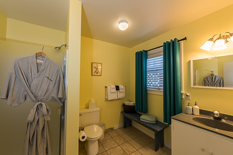 A bright and cheerful bathroom with a yellow wall, a white vanity, a toilet, and a shower. A robe hangs on the door and a window with blue curtains provides natural light.