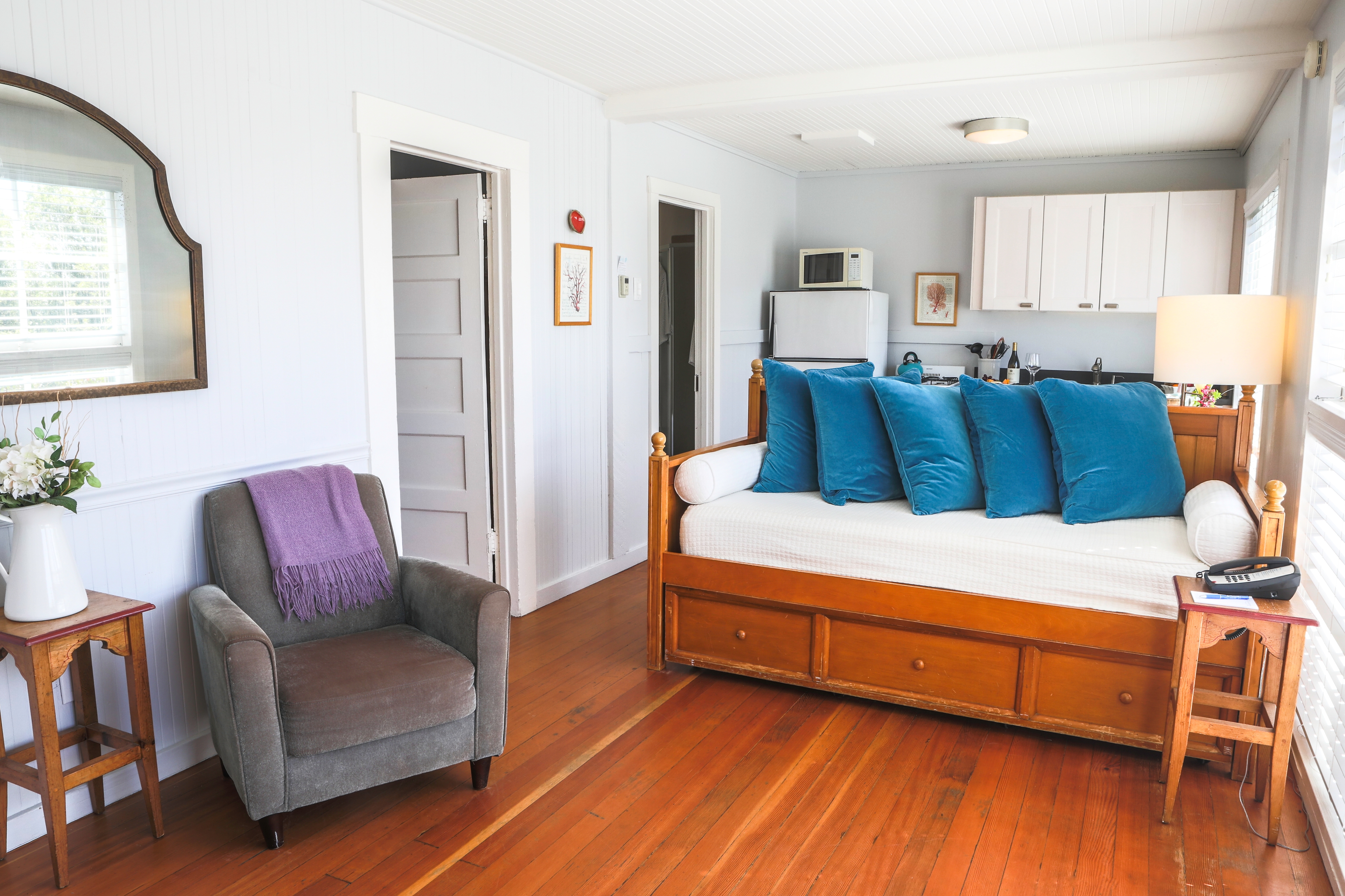 A cozy living area with a wooden daybed, a comfortable armchair, and a small kitchenette, featuring a whitewashed wall and hardwood floors.