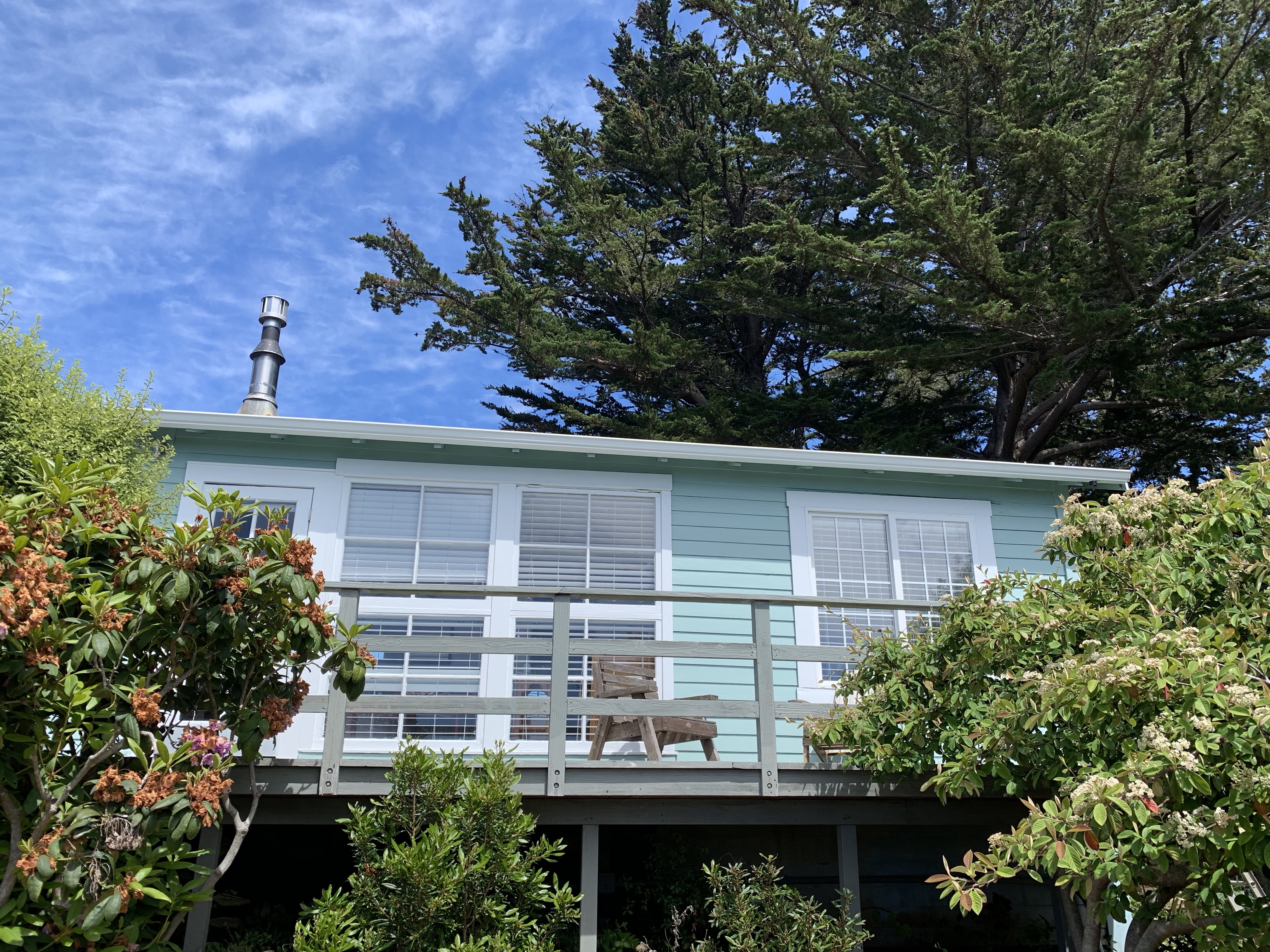 A cozy cottage with a balcony and large windows, surrounded by trees and greenery.