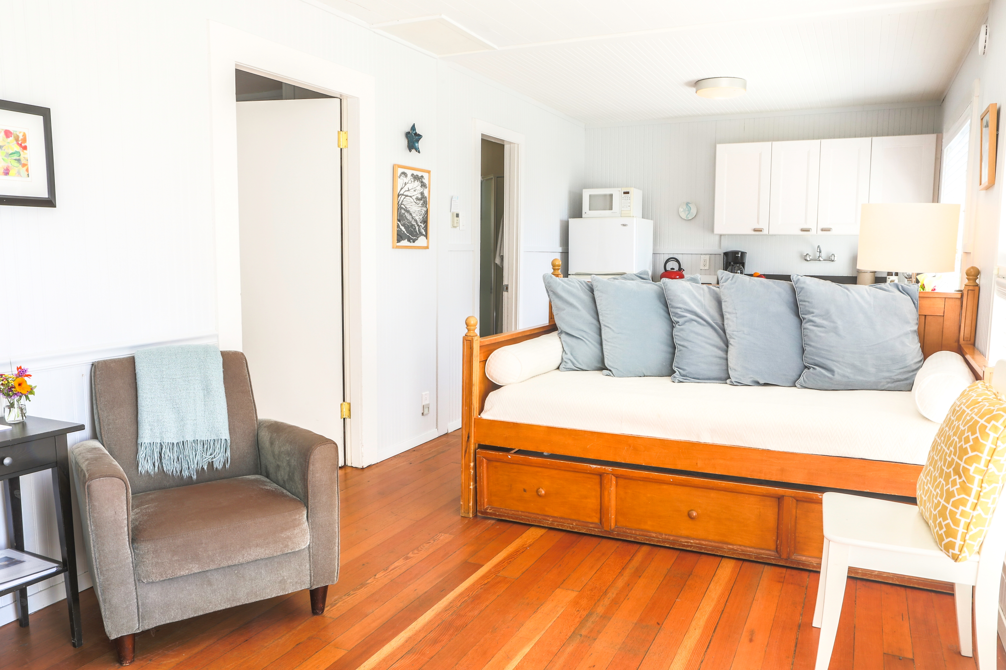 A bright and airy room with a wooden daybed, a comfortable armchair, and a small kitchenette with a microwave and a refrigerator.