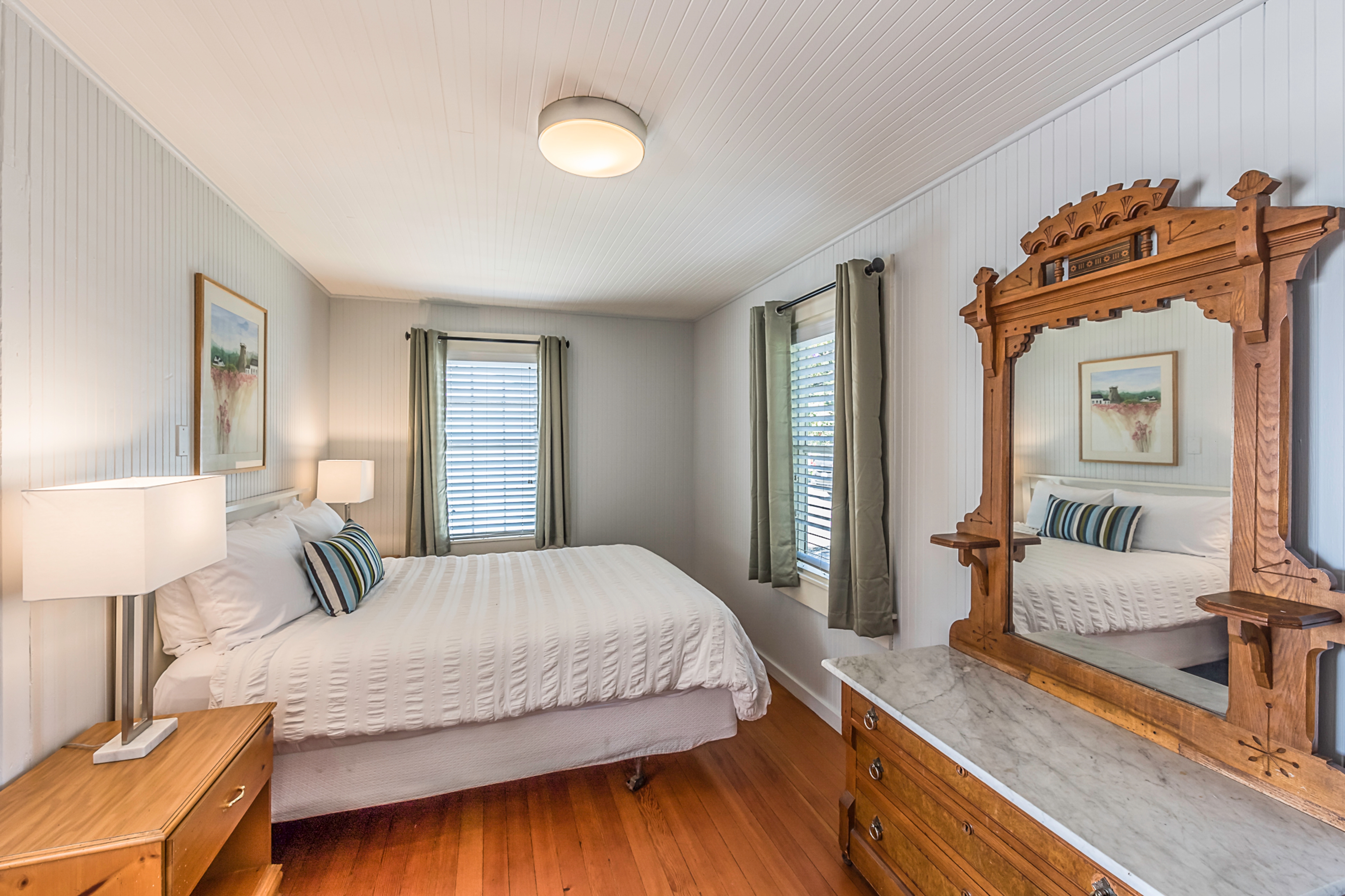 A cozy bedroom with a white bed, two nightstands, a dresser with an ornate wooden mirror, and two windows with curtains..