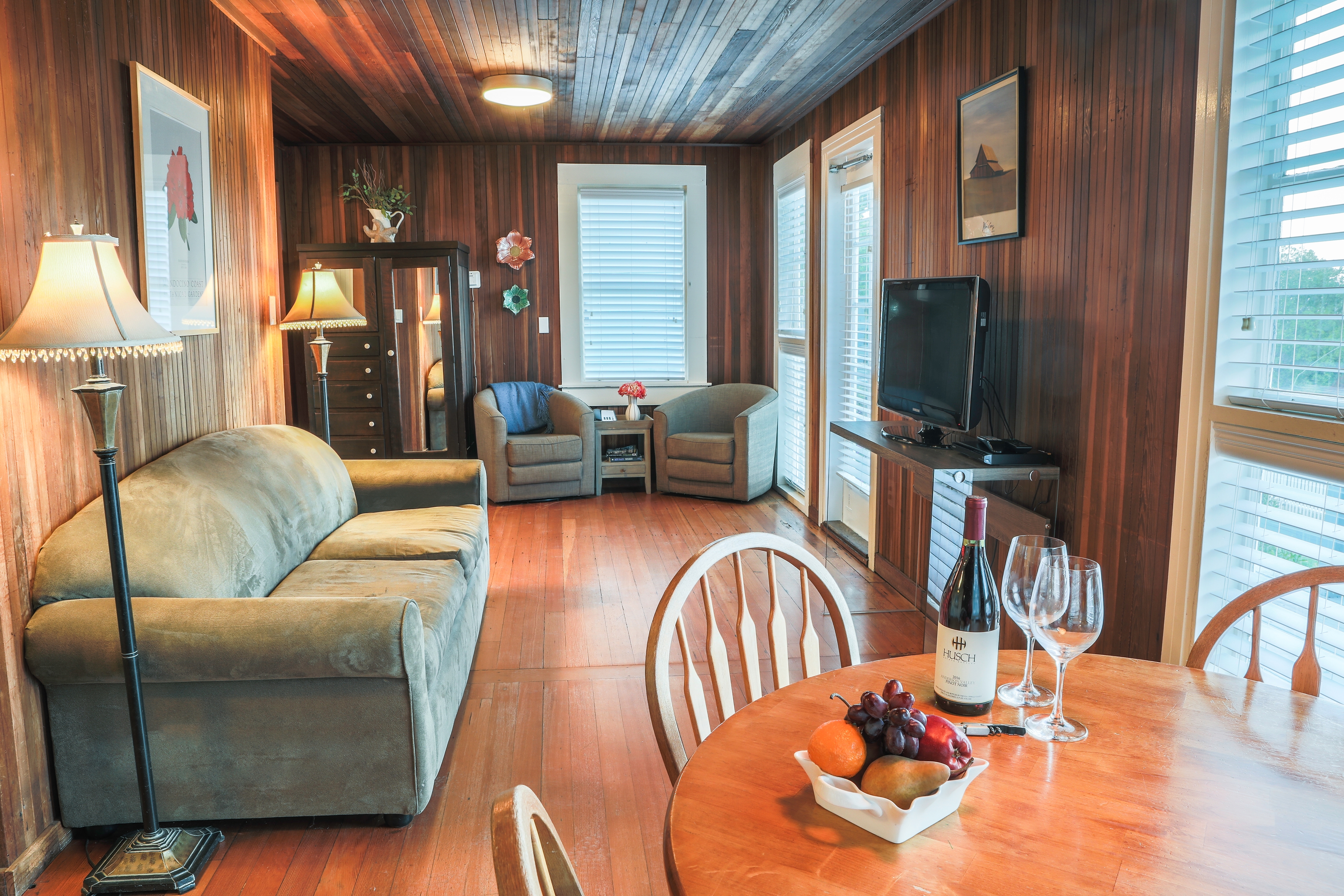 A wood-paneled room with a gray sofa, two armchairs, a dining table, and a view of the outdoors through large windows.