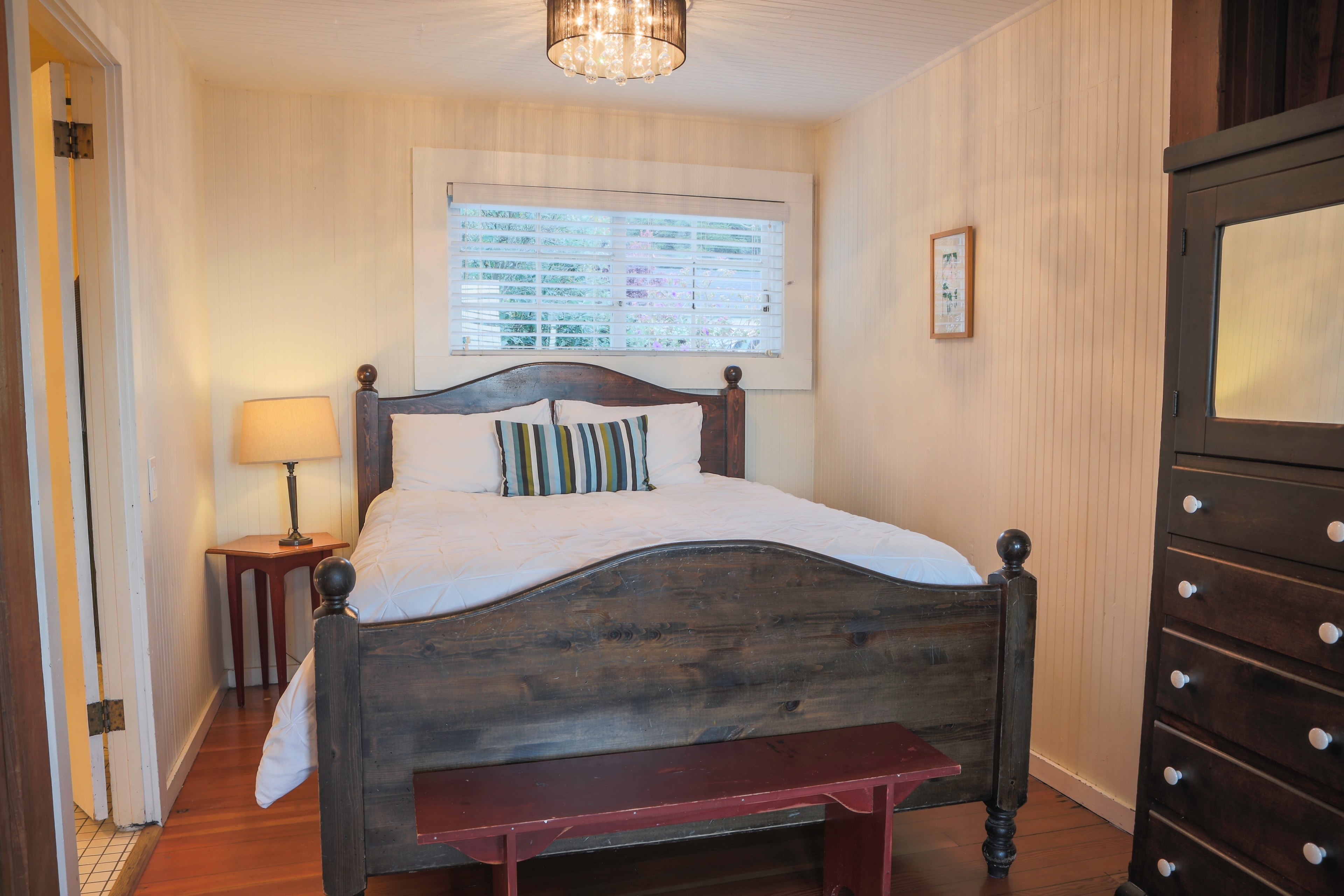 A cozy bedroom with a wooden bed, a nightstand, hardwood floors, and a window with a view of the outdoors.