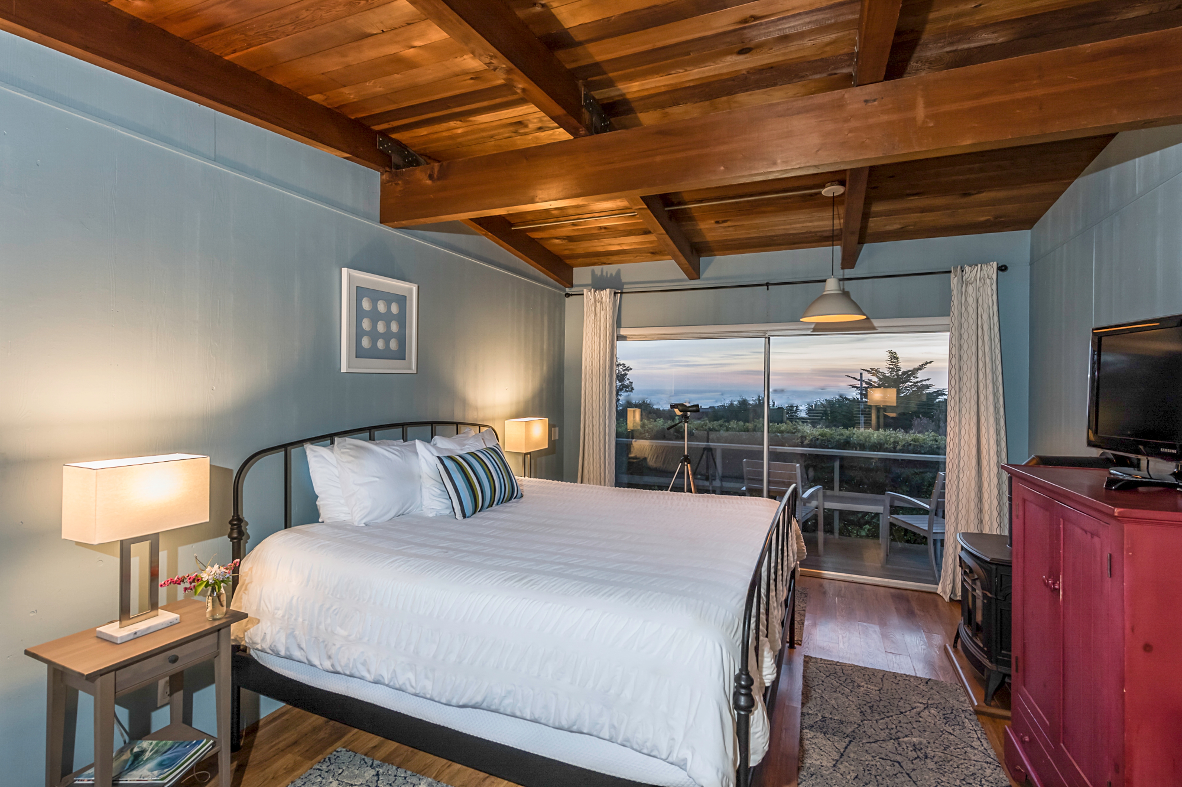 Van Damme Bedroom with a king-sized bed, a wood-burning stove, and a view of the outdoors through large sliding glass doors, featuring a wood-beamed ceiling.