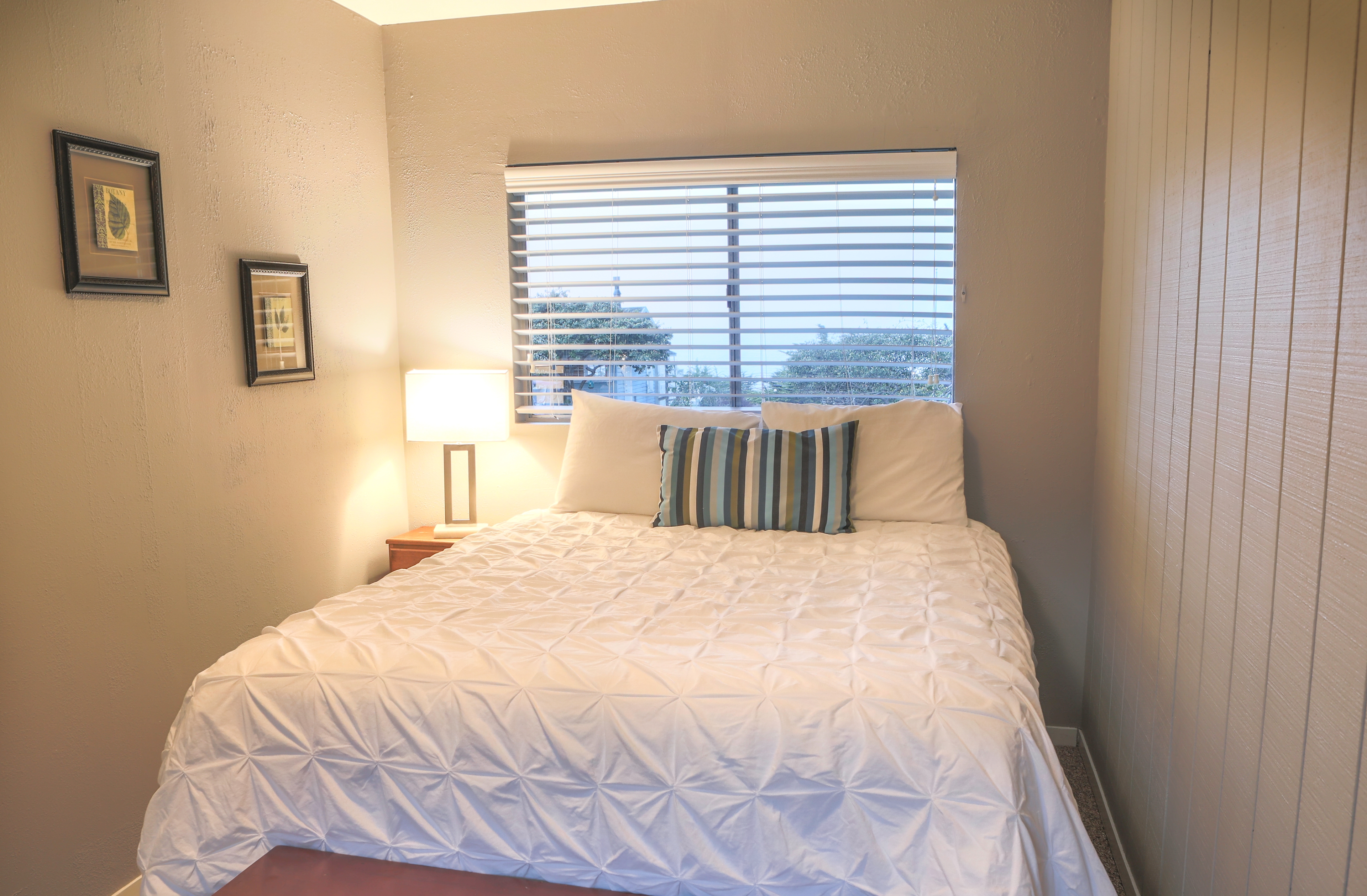 A simple bedroom with a white bed, a nightstand, and a window, featuring a light-colored wall and hardwood floors.