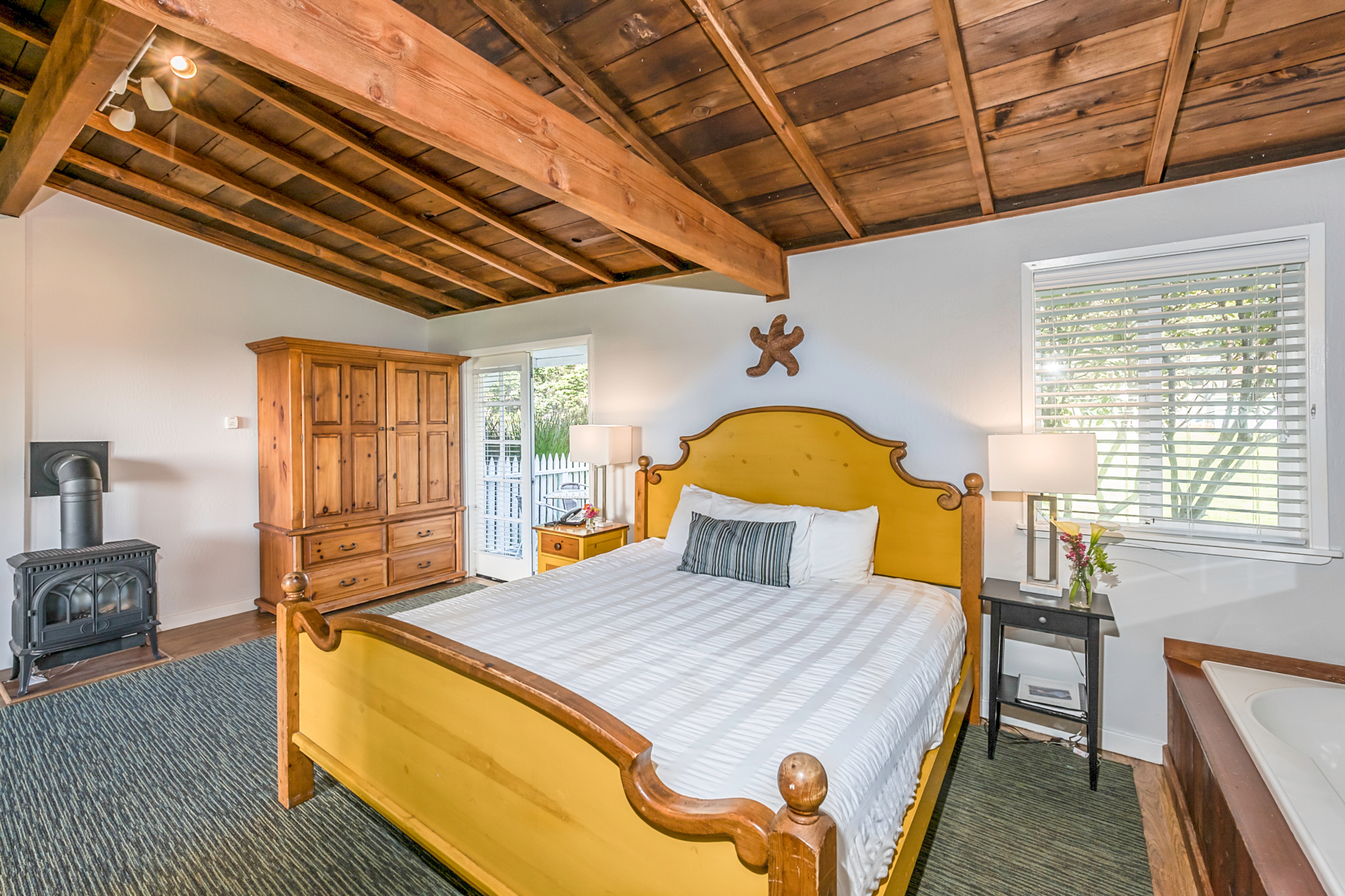 A spacious bedroom with a yellow bed, a wood-burning stove, featuring a wood-beamed ceiling, and a view of the outdoors through a window.