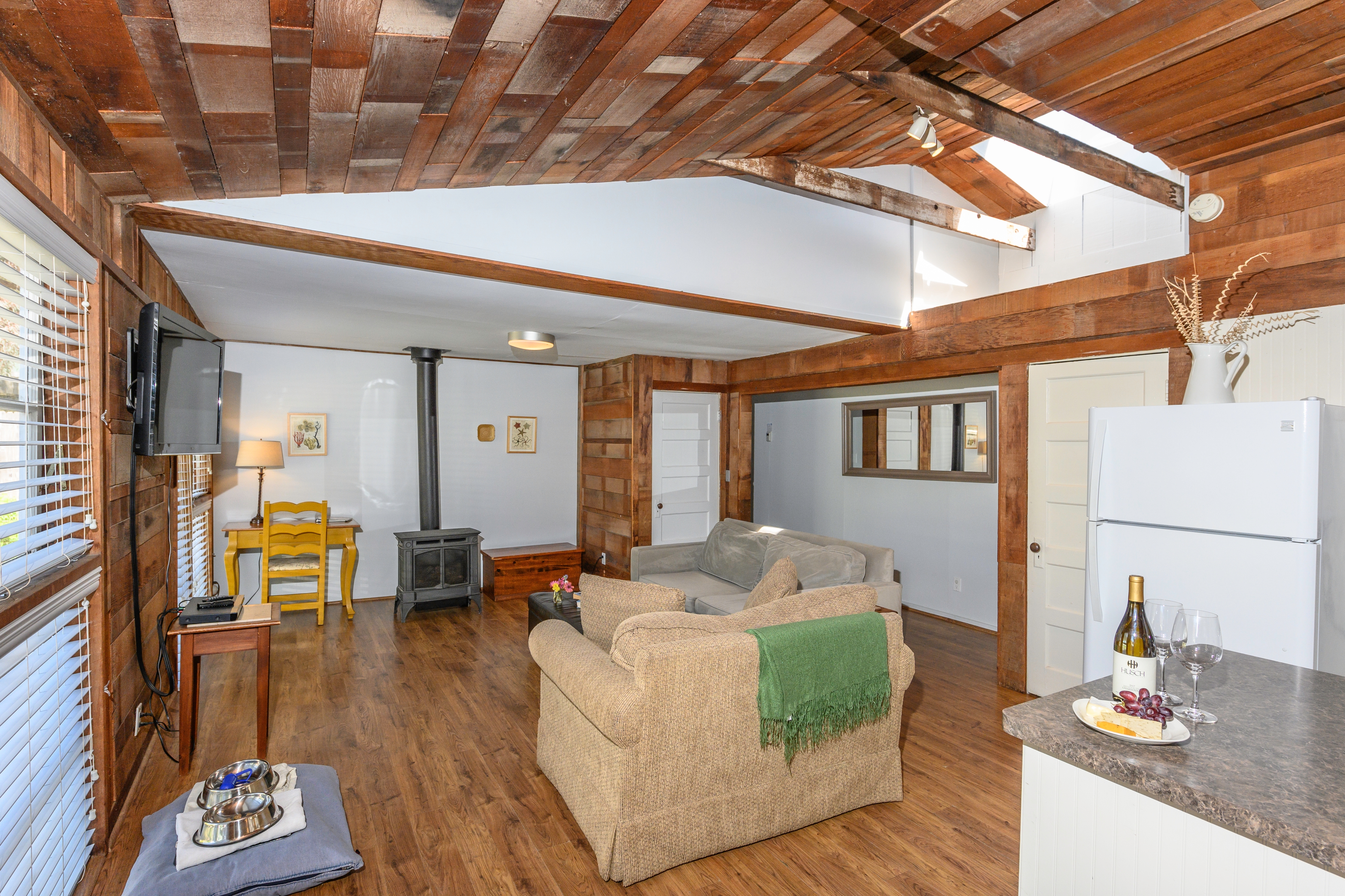 A rustic living space with wood-paneled walls and a wood-beamed ceiling, offering plenty of seating and a view of a bedroom.