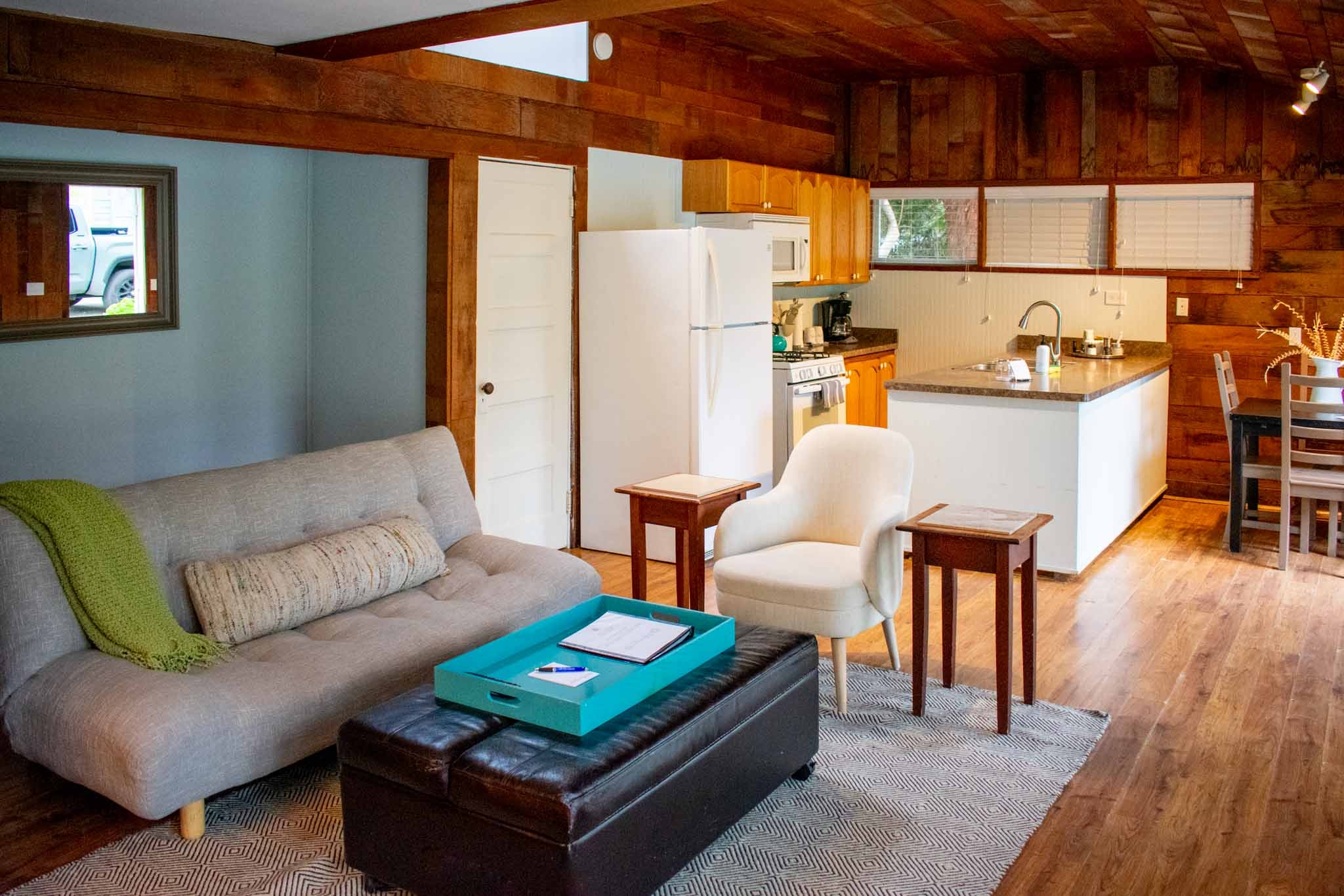 An open floor rustic kitchen with wood-paneled walls and a wood-beamed ceiling, featuring a kitchen island, a dining table, and a view of the outdoors through a window.