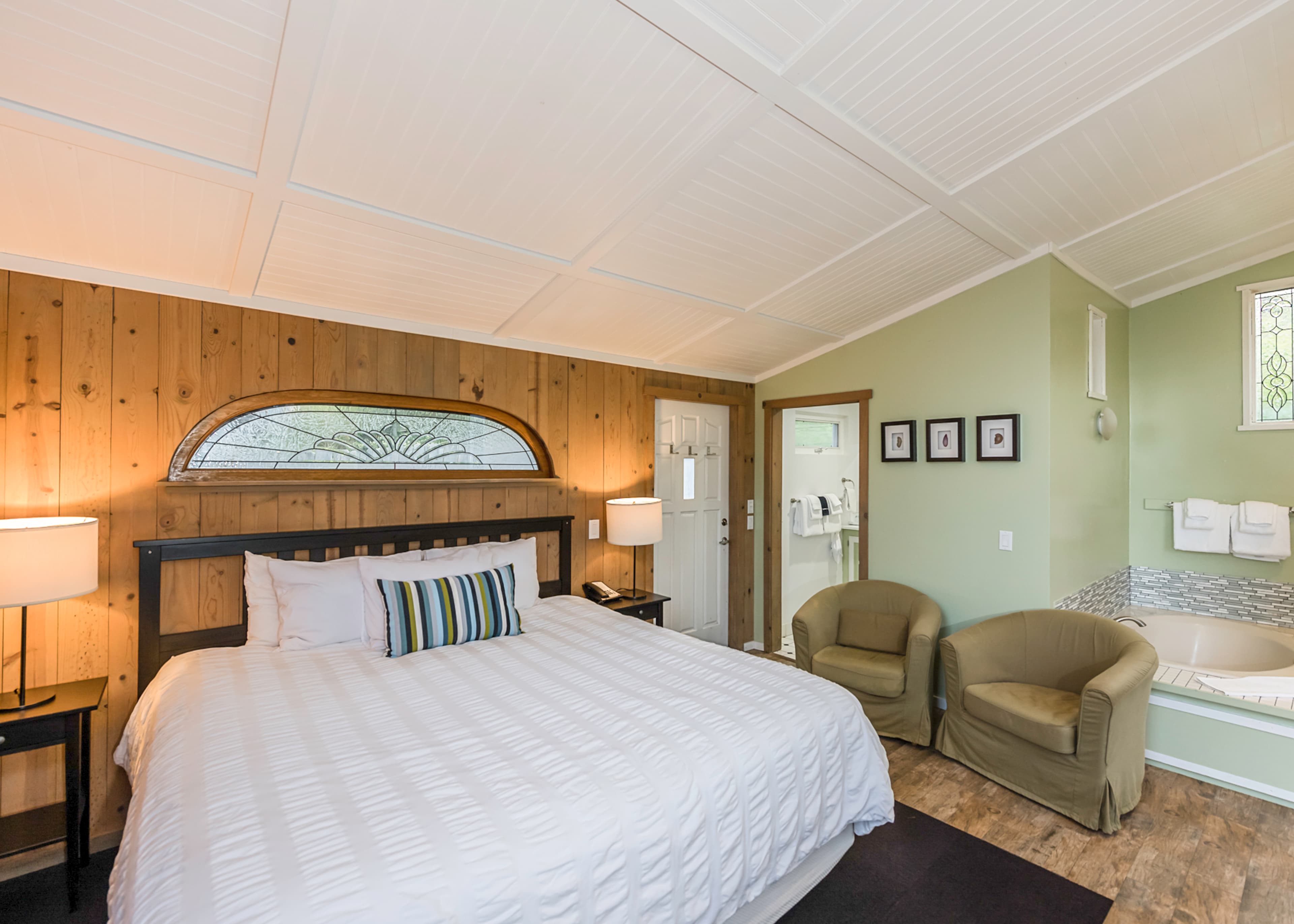 A spacious bedroom with a king-sized bed, a green armchair, and a bathtub with a view, featuring wood-paneled walls and a vaulted ceiling.