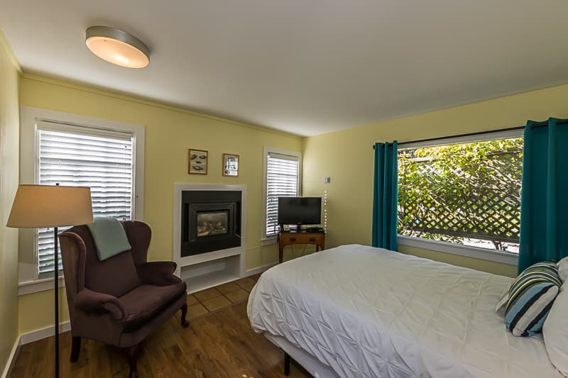 A bright and airy bedroom with a queen-sized bed, a brown armchair, a fireplace, and a large window with a view of the outdoors.