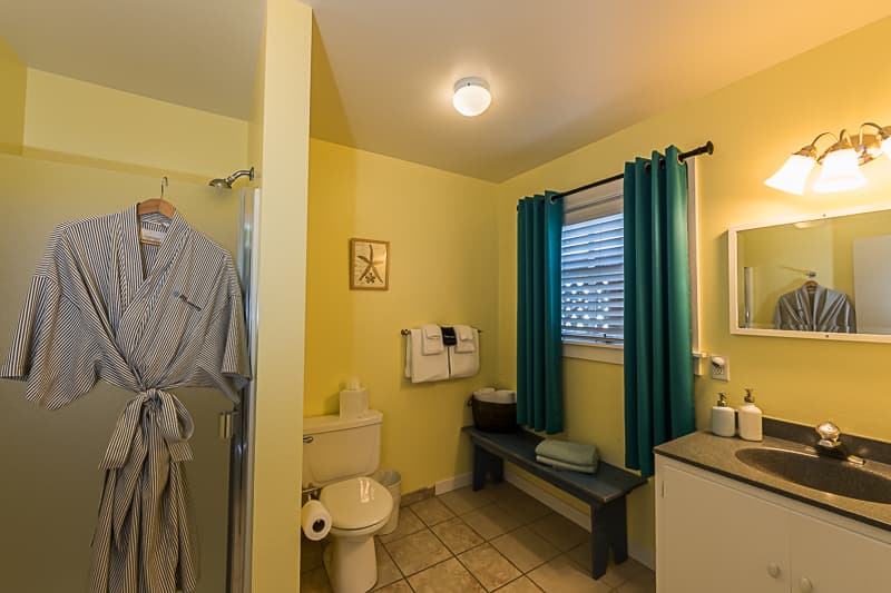 A bright and cheerful bathroom with a yellow wall, a white vanity, a toilet, and a shower. A robe hangs on the door and a window with blue curtains provides natural light.