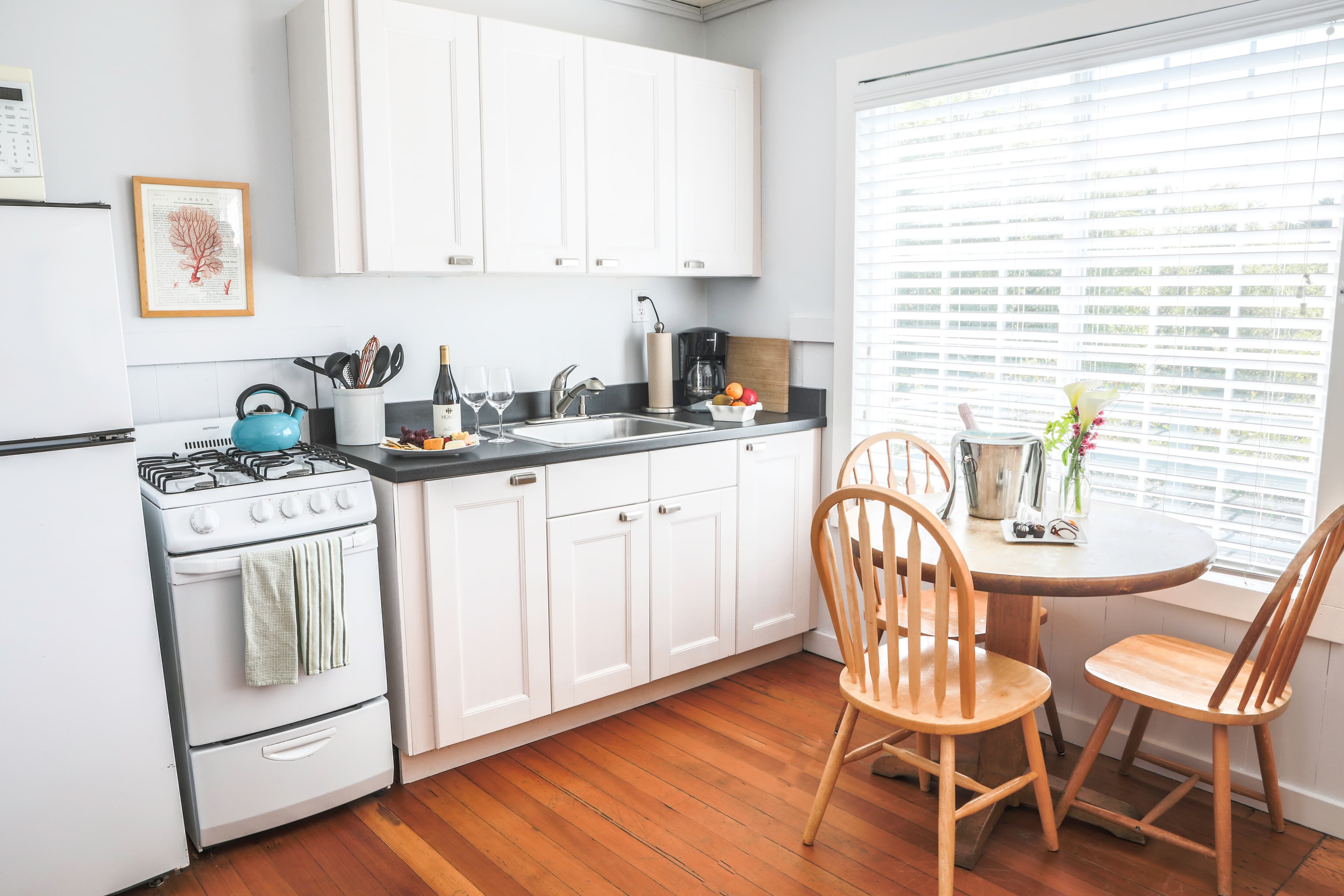 A small, well-equipped kitchenette with white cabinets, a stove, a refrigerator, and a sink. A small dining table with two chairs is set up next to a window.