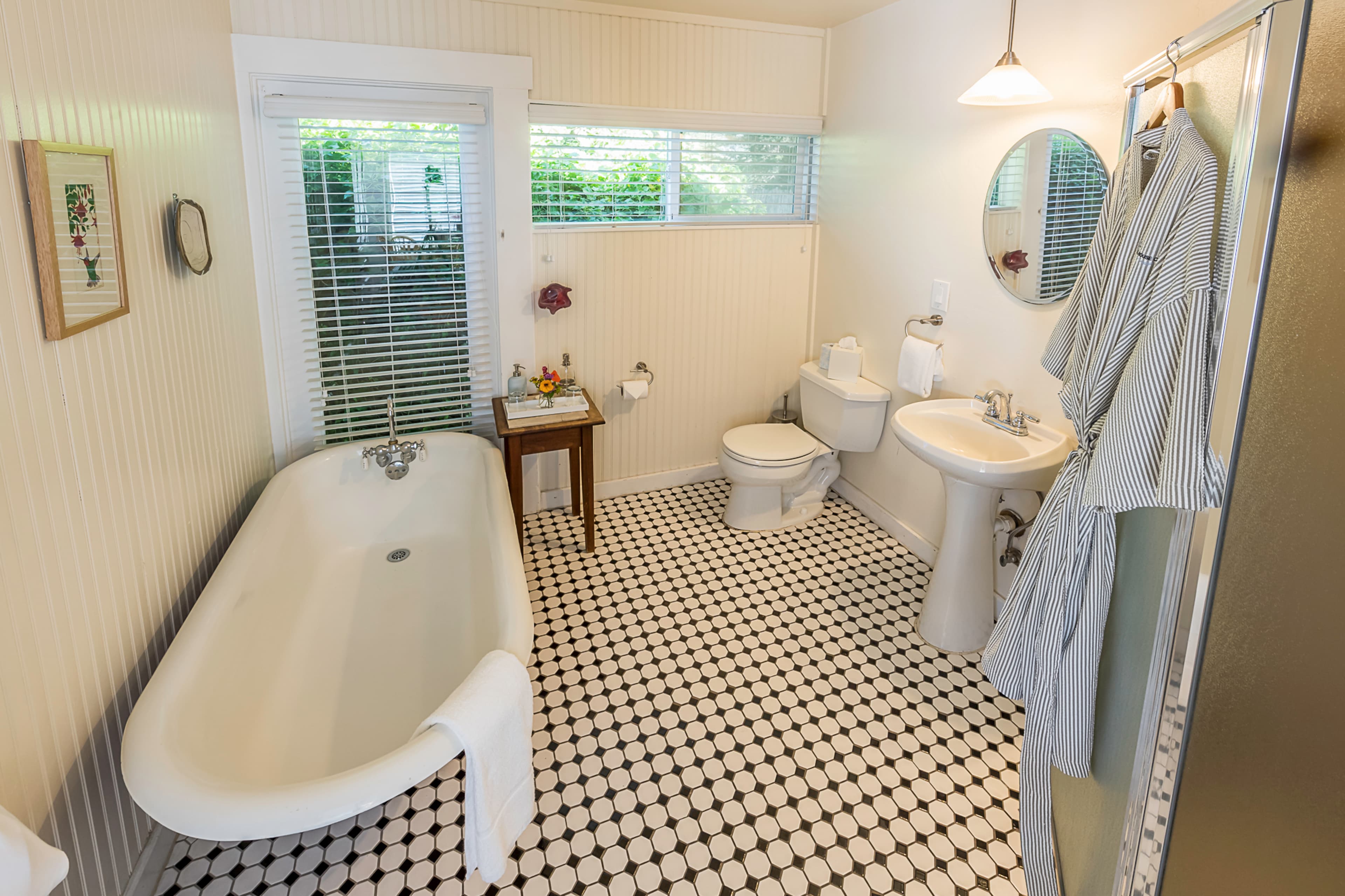 A classic bathroom with a vintage clawfoot bathtub, a toilet, and a sink. Two plush robes hang on a hook on the wall, and a window provides natural light.