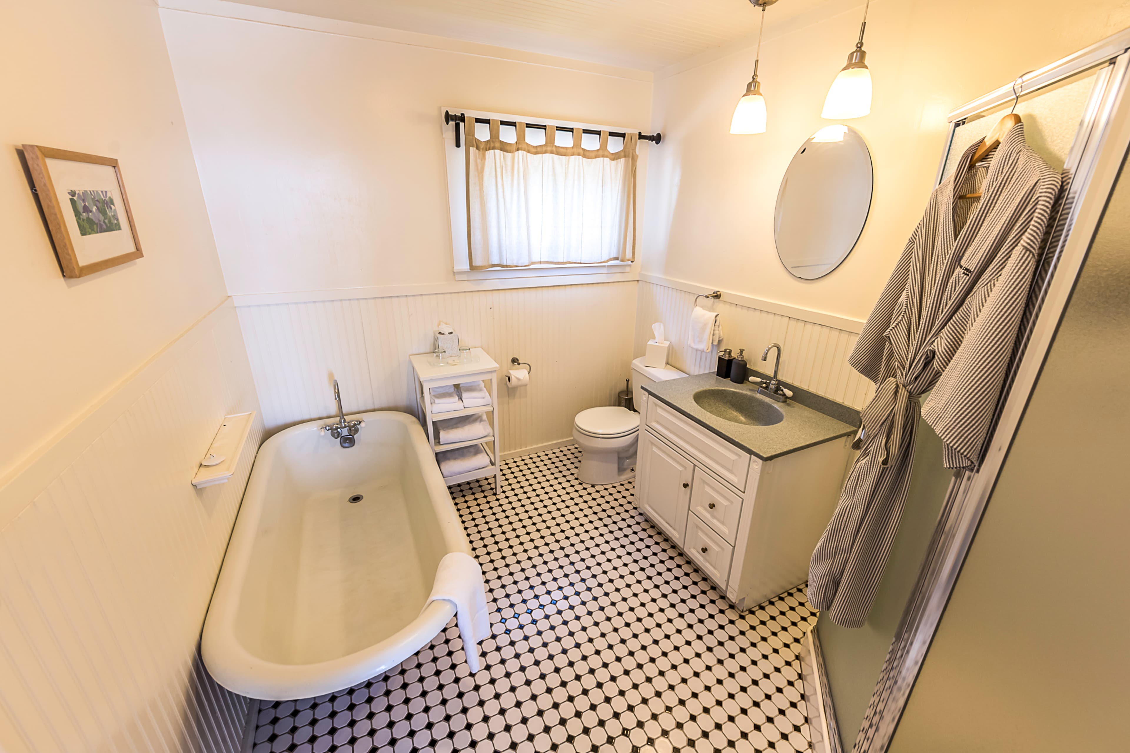 A classic bathroom with a vintage clawfoot bathtub, a toilet, and a sink. Two plush robes hang on a hook on the wall, and a window provides natural light.