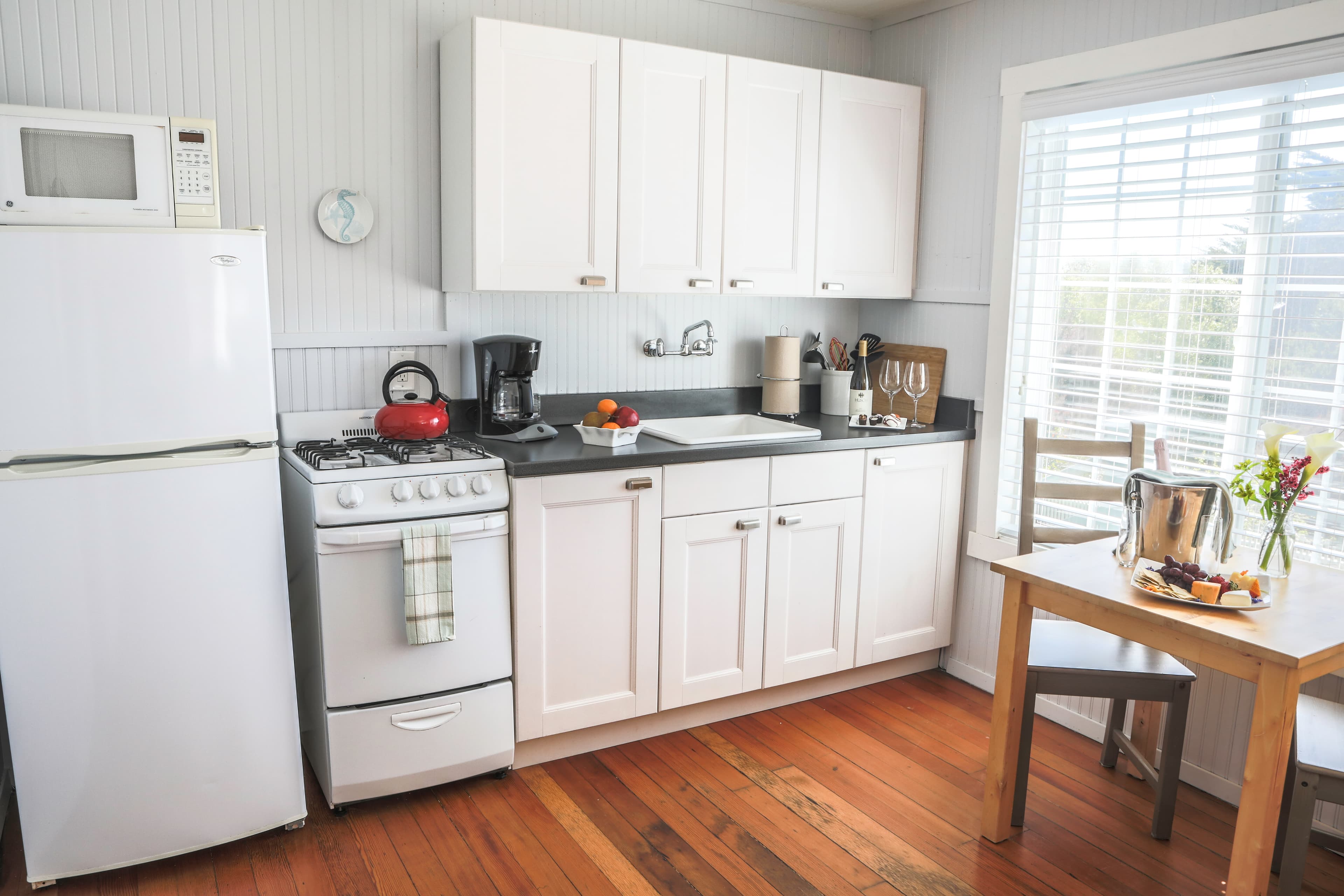 A small, well-equipped kitchenette with white cabinets, a stove, a refrigerator, and a sink. A small dining table with two chairs is set up next to a window.