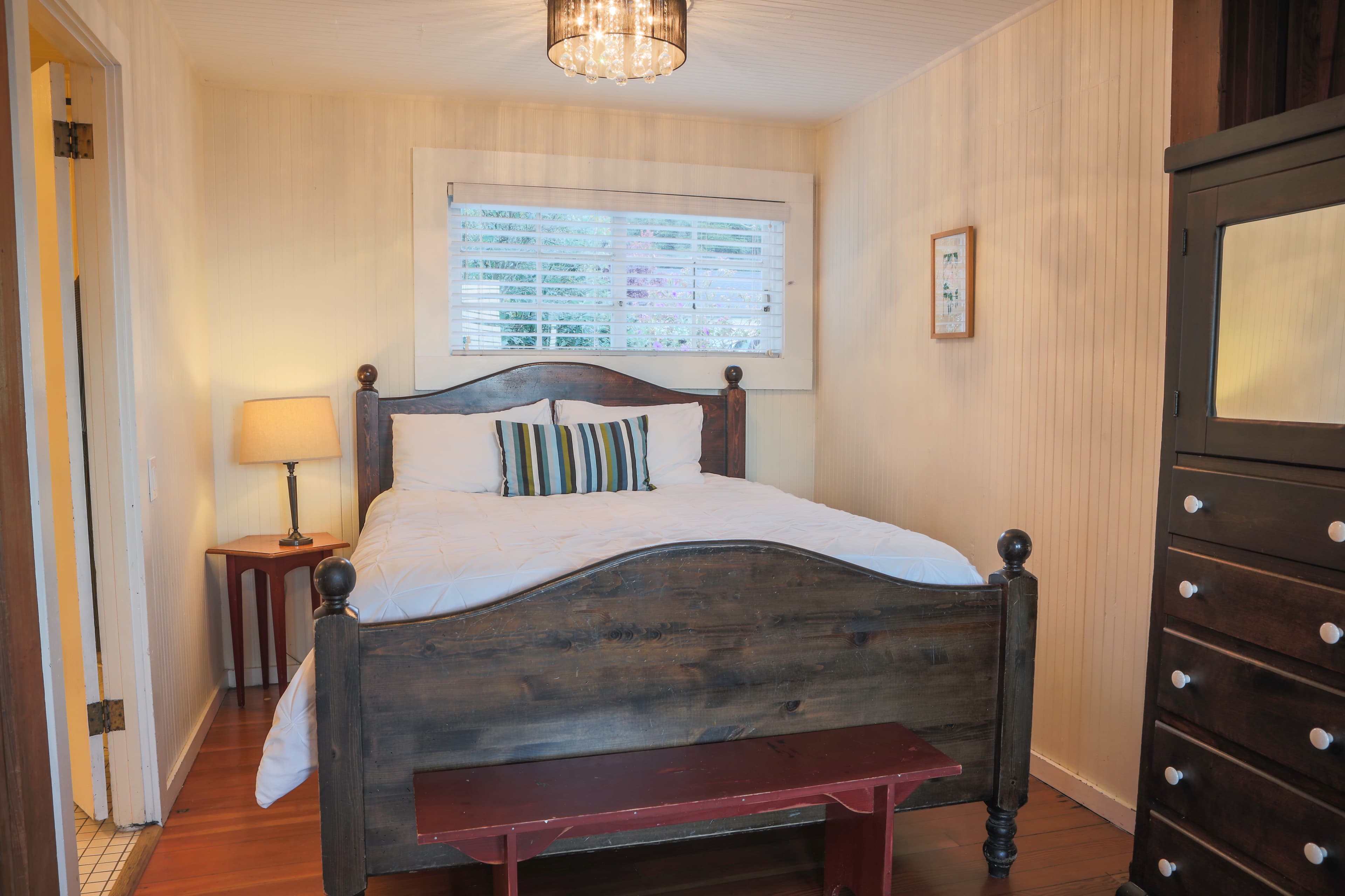 A cozy bedroom with a wooden bed, a nightstand, hardwood floors, and a window with a view of the outdoors.