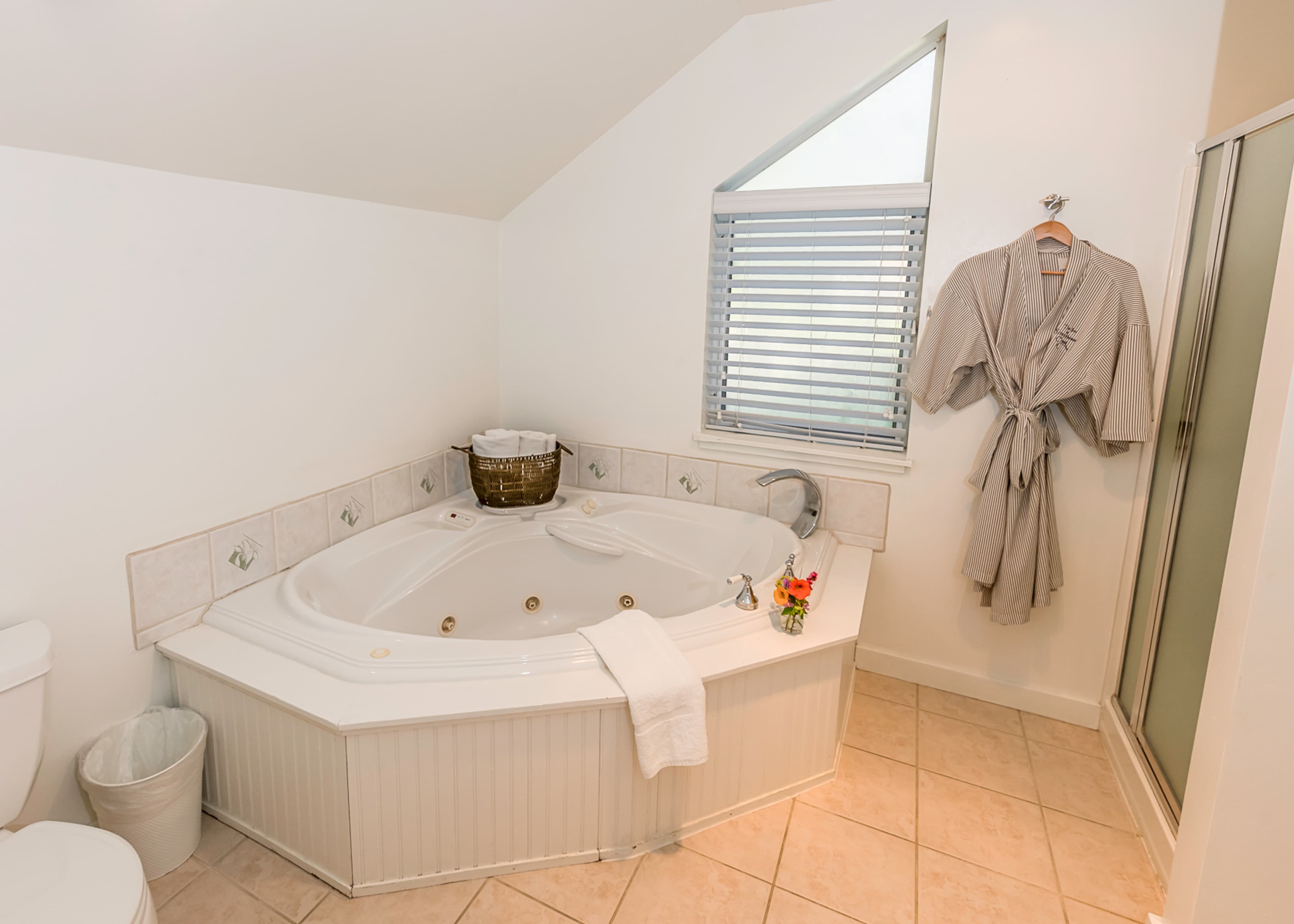 A luxurious bathroom with a large, corner jetted tub, a toilet, and a shower. A robe hangs on a hook on the wall, and a window provides natural light.