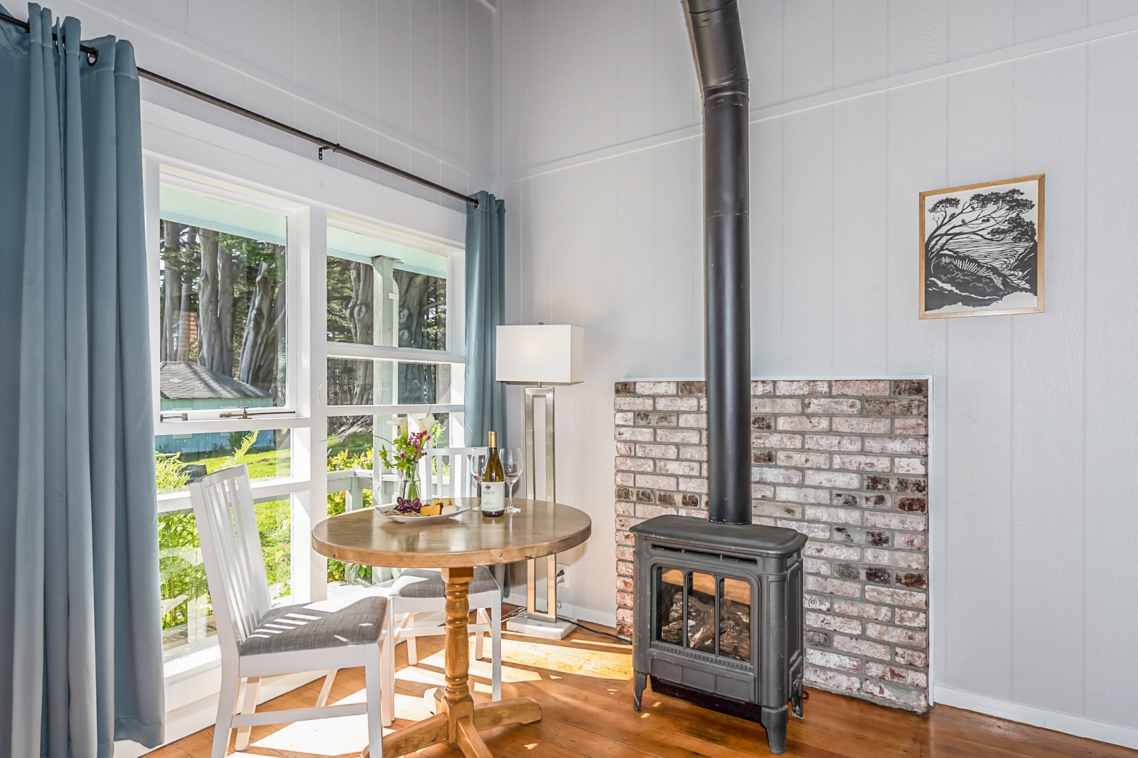 A cozy room with a wood-burning stove, a small dining table, and large windows offering a view of the outdoors.