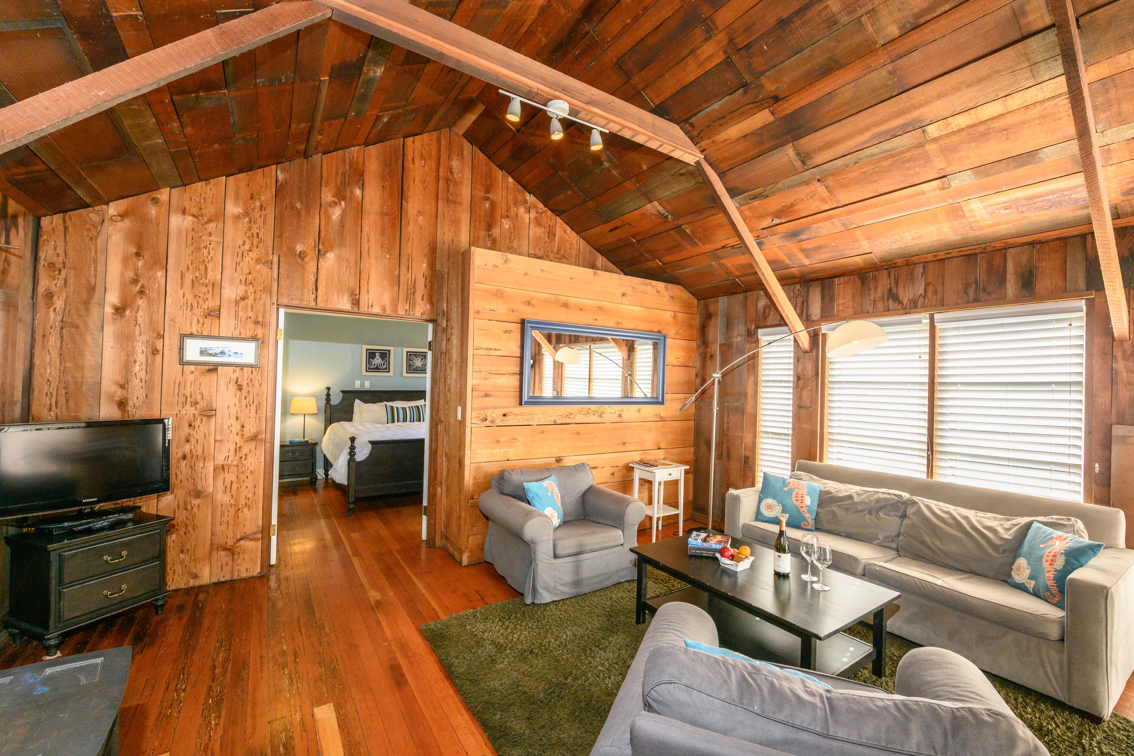 A rustic living space with wood-paneled walls and a wood-beamed ceiling, offering plenty of seating and a view of a bedroom.