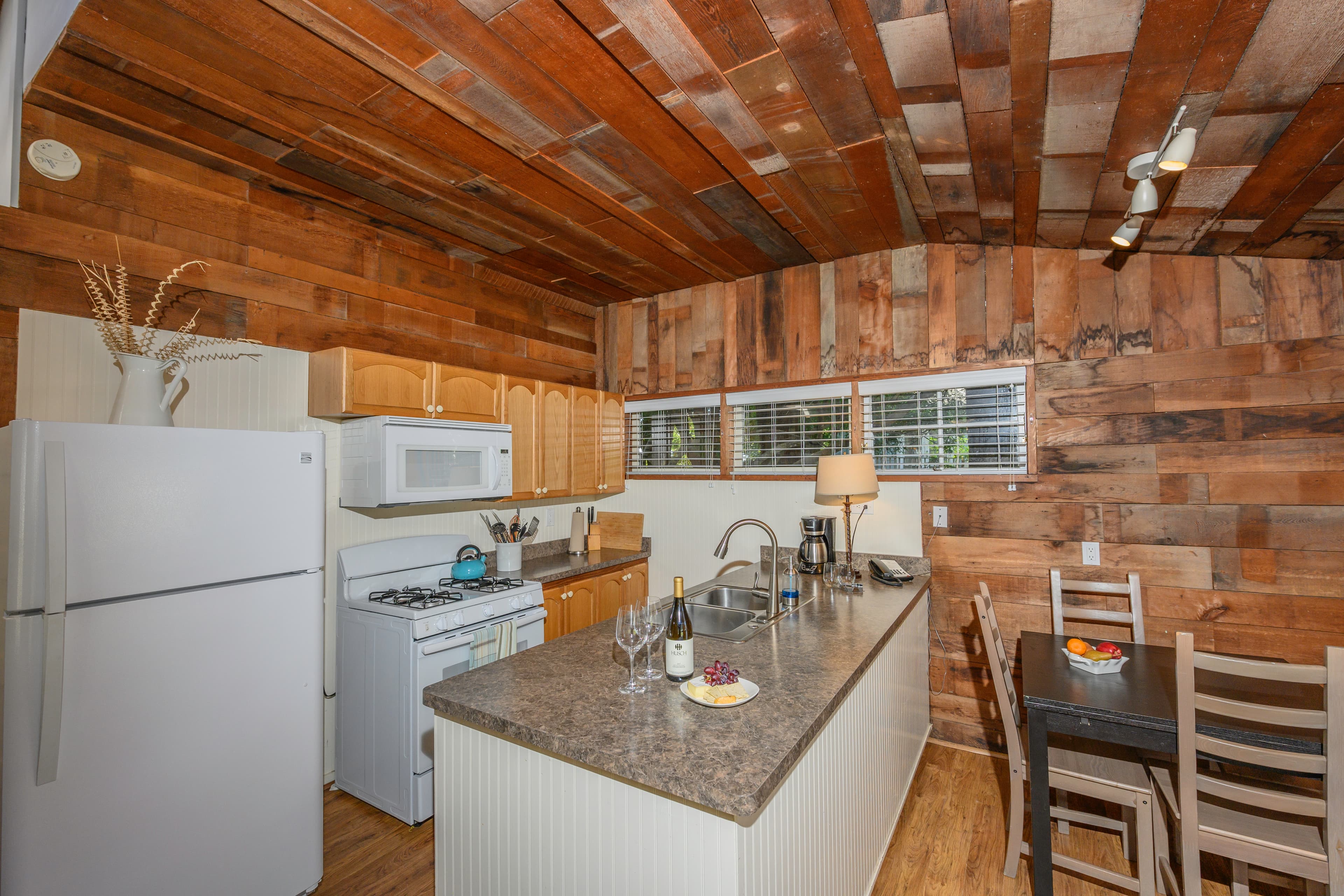 A rustic kitchen with wood-paneled walls and a wood-beamed ceiling, featuring a kitchen island, a dining table, and a view of the outdoors through a window.