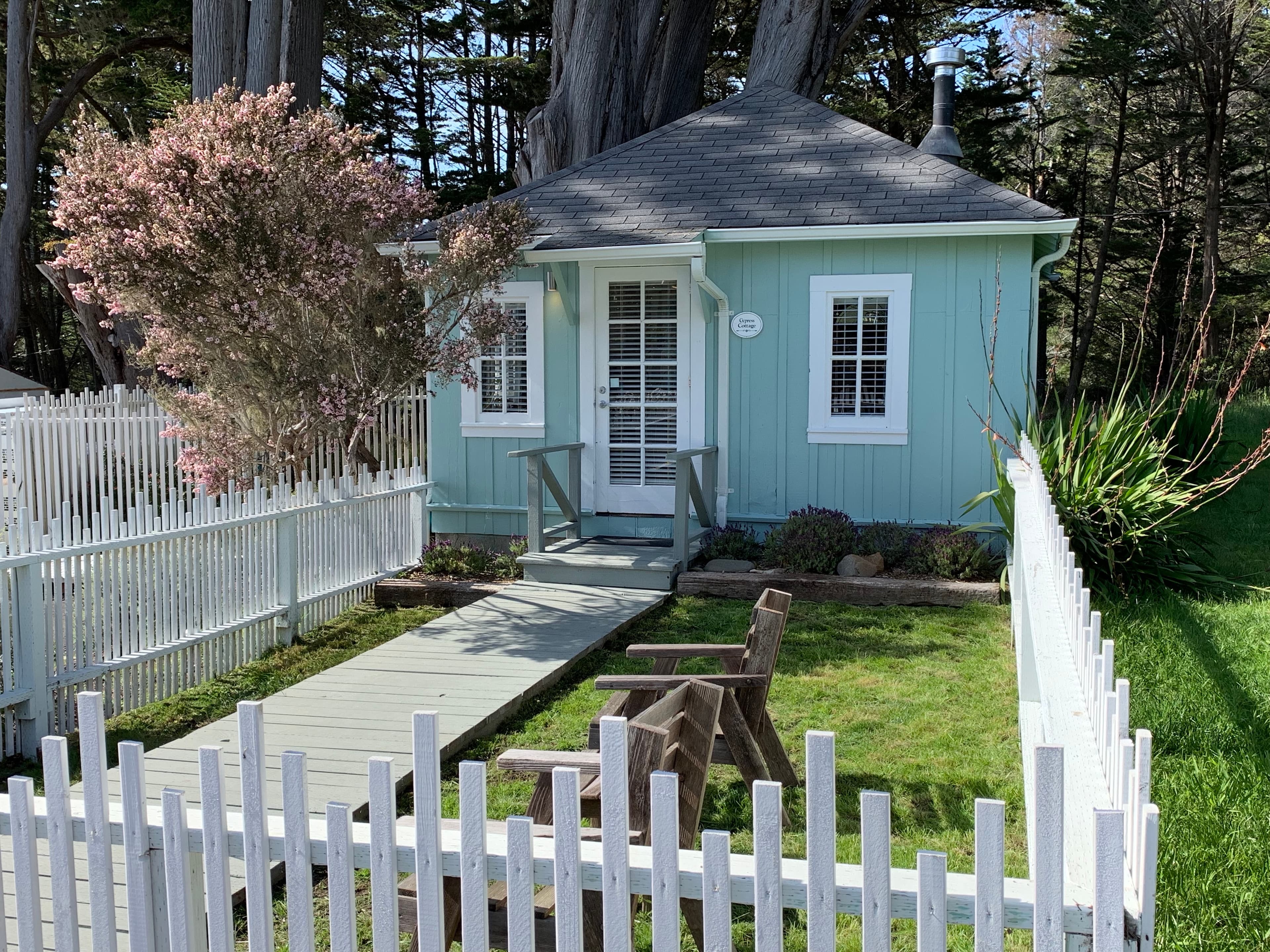 A small, light blue cottage with a white picket fence and a walkway leading to the front door. There is a bench and a small table in the front yard.