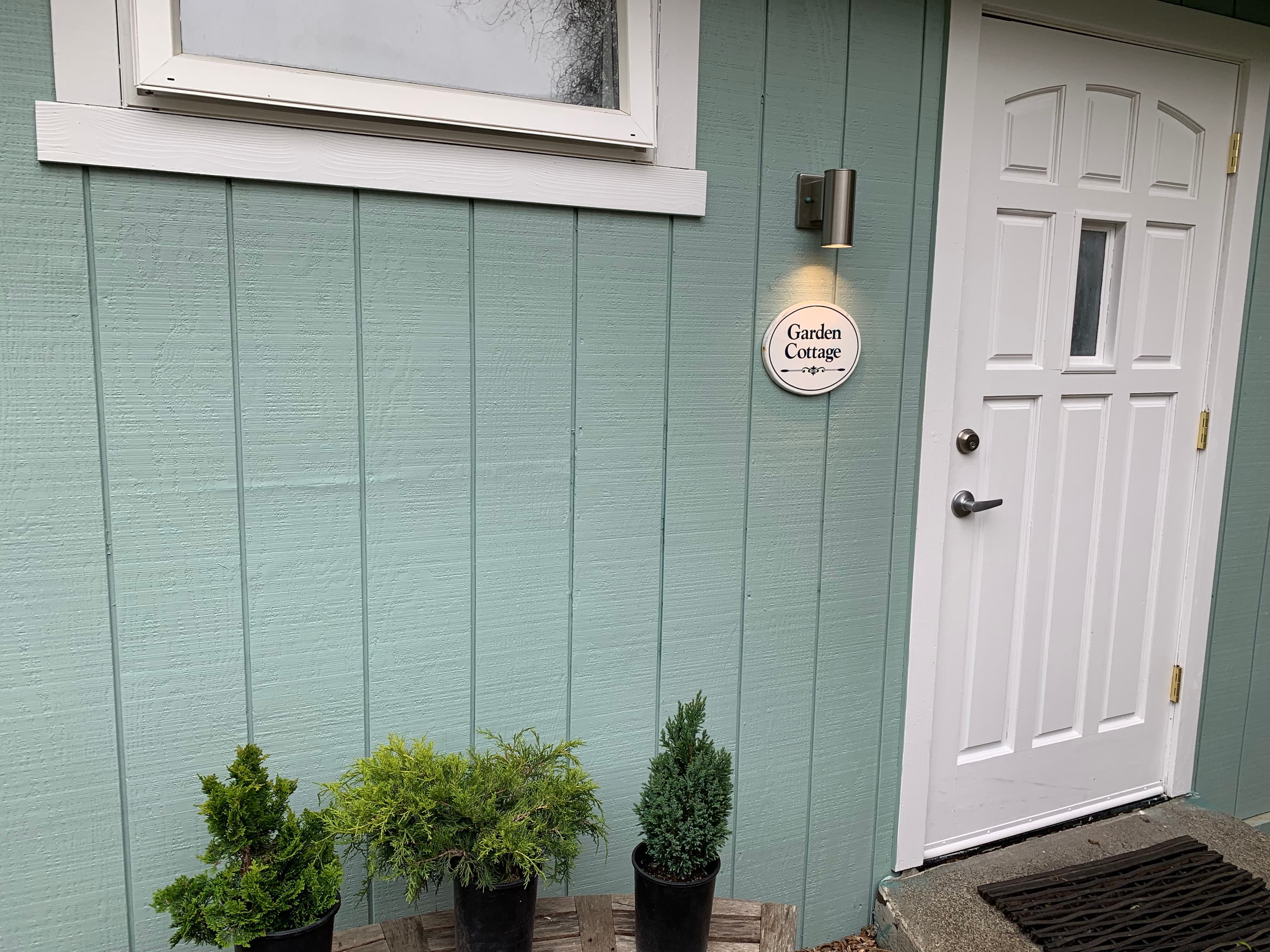 A white door with a small window, flanked by potted plants. A welcome sign is attached to the wall beside the door.
