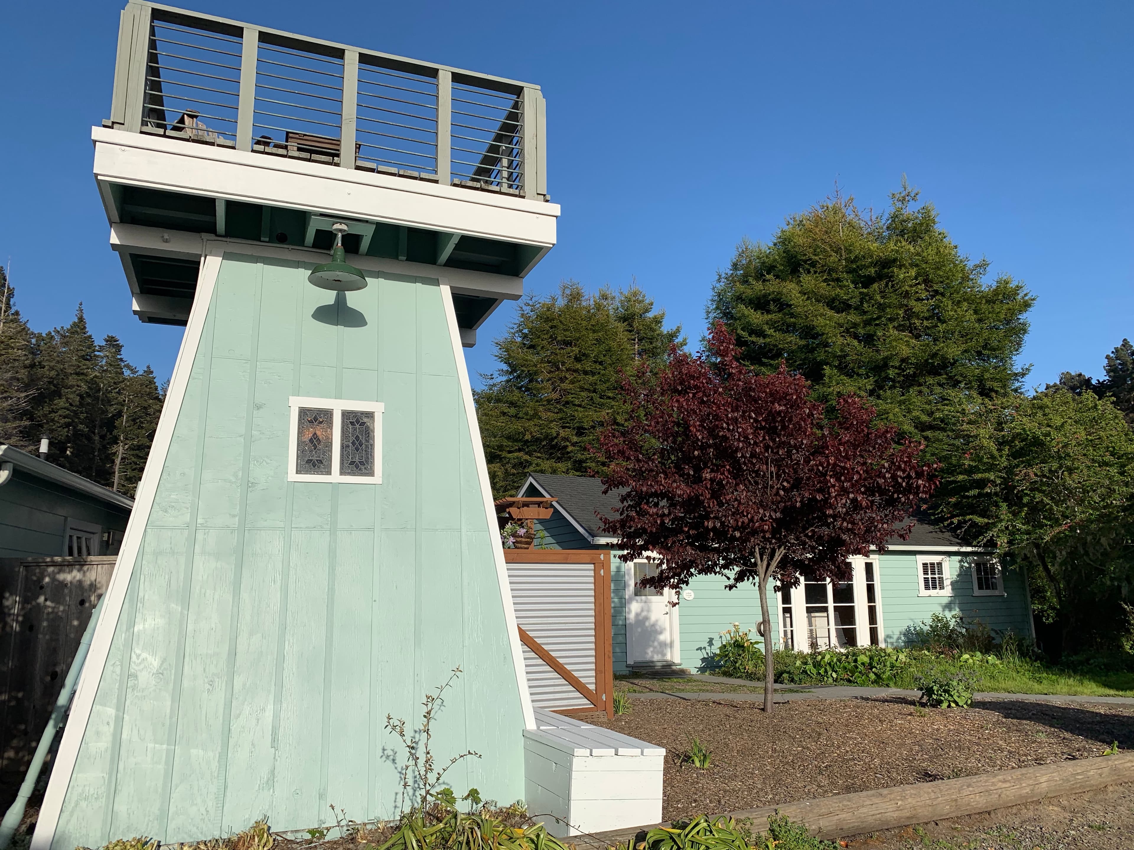 A unique two-story house with a tower and a rooftop deck, surrounded by trees and greenery.