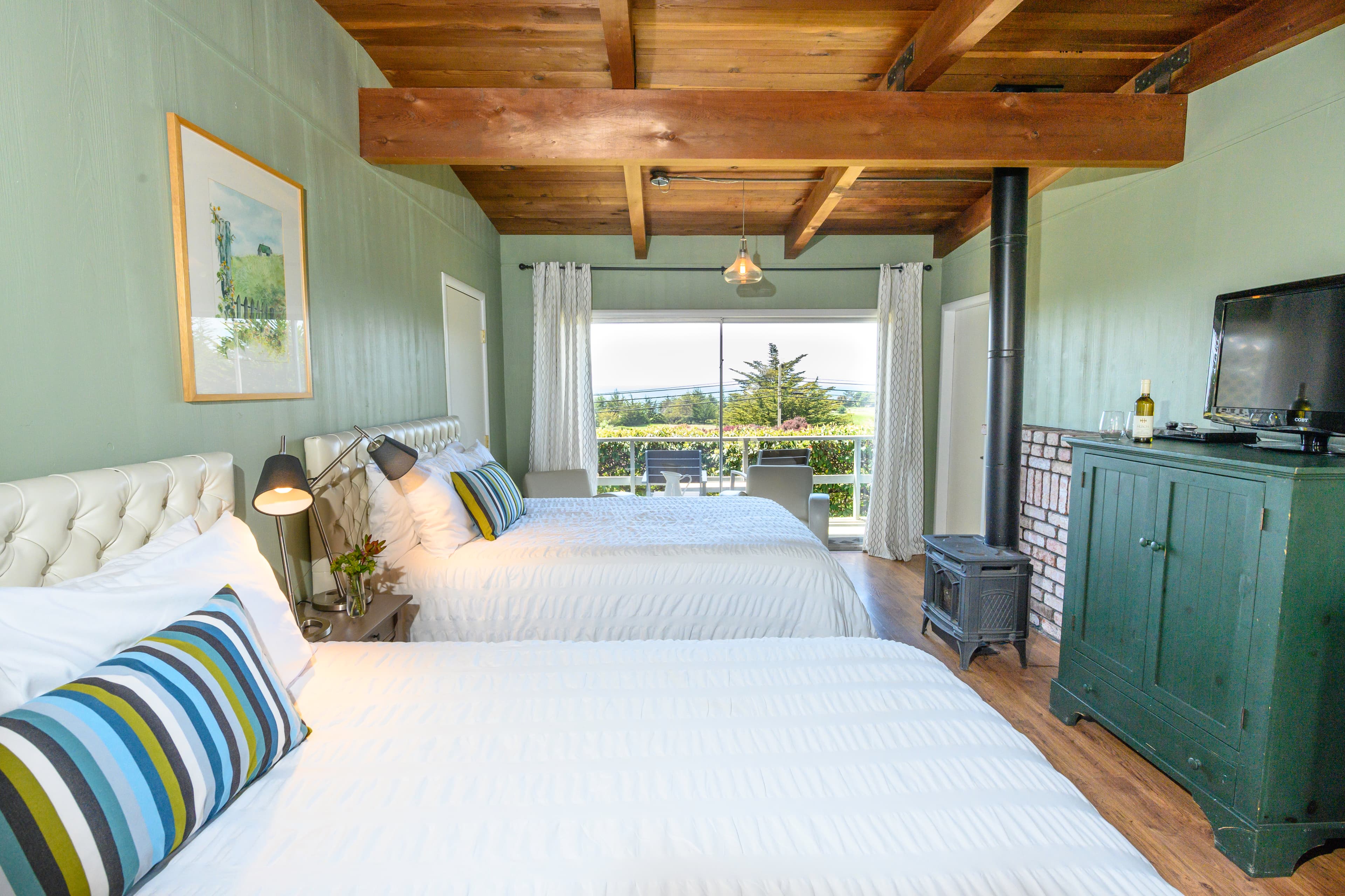 A cozy bedroom with two twin beds, a wood-burning stove, and a view of the outdoors through large sliding glass doors.