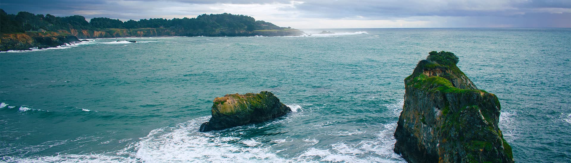 Coastal scene with rocky outcrops and waves crashing against the shore under a cloudy sky.