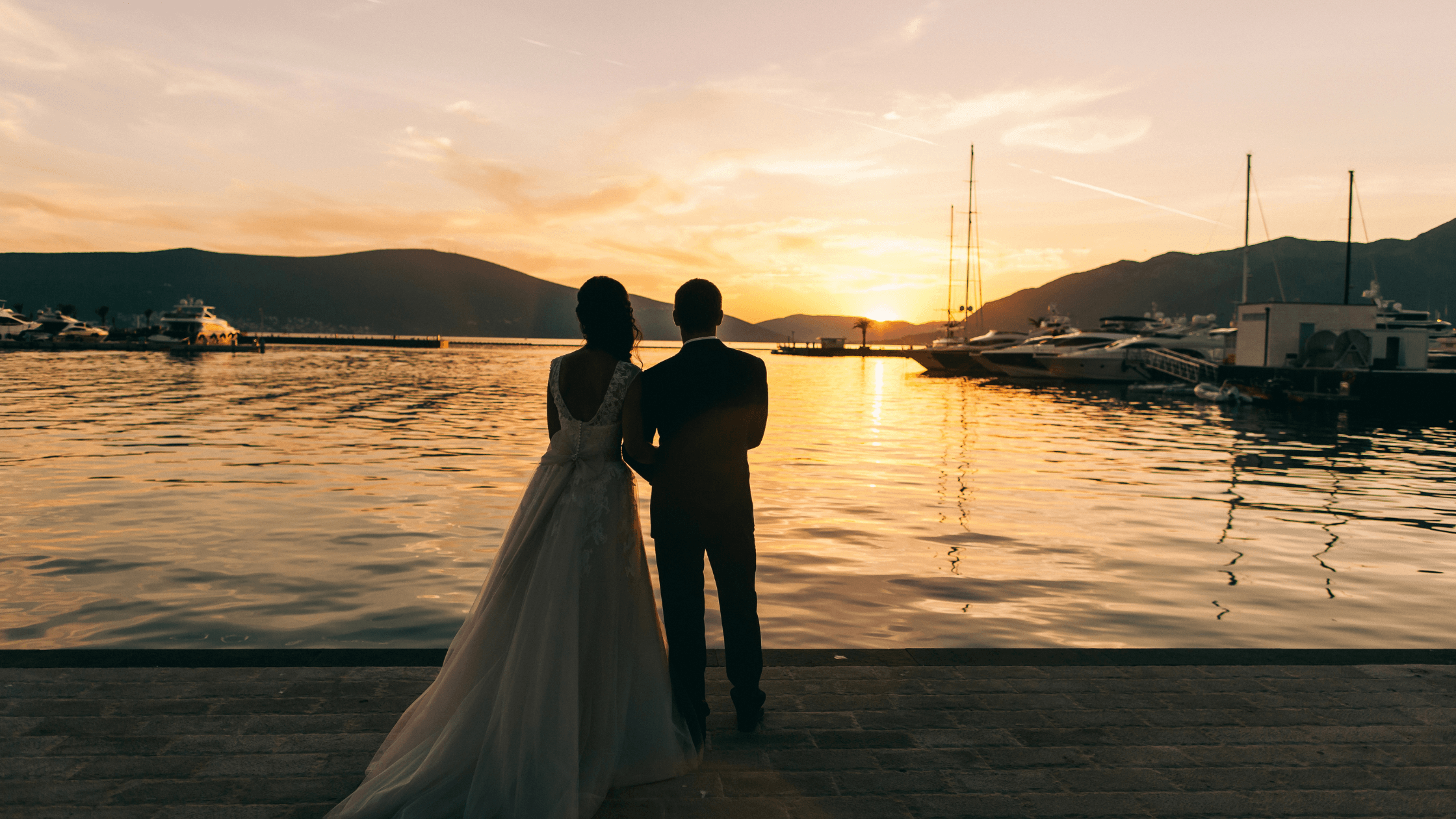 A couple stands together by the water, silhouetted against a sunset. A couple stands together by the water, silhouetted against a sunset.