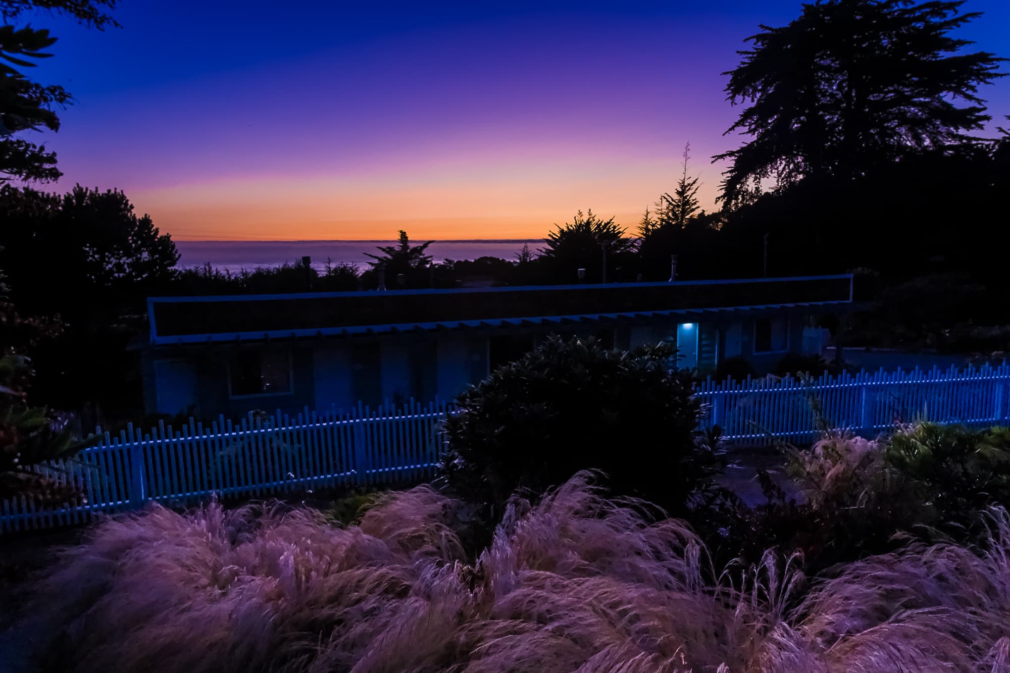 A tranquil coastal scene at dusk with a colorful sky and a white fence surrounding a building.