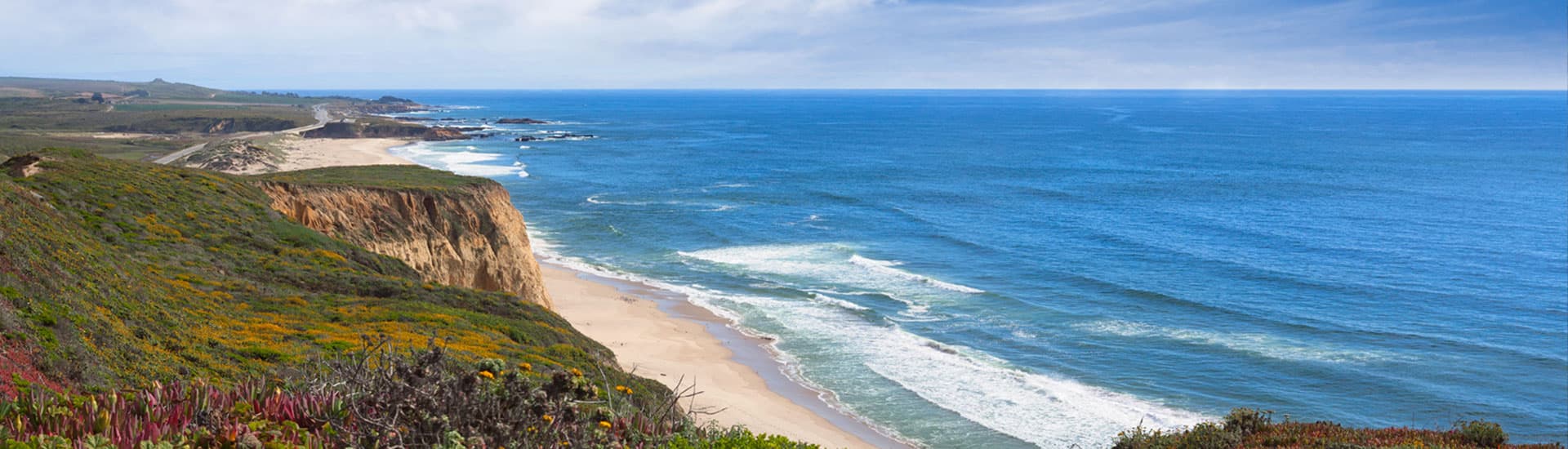 A scenic coastal view featuring cliffs, sandy beaches, and vast ocean waves under a partly cloudy sky.