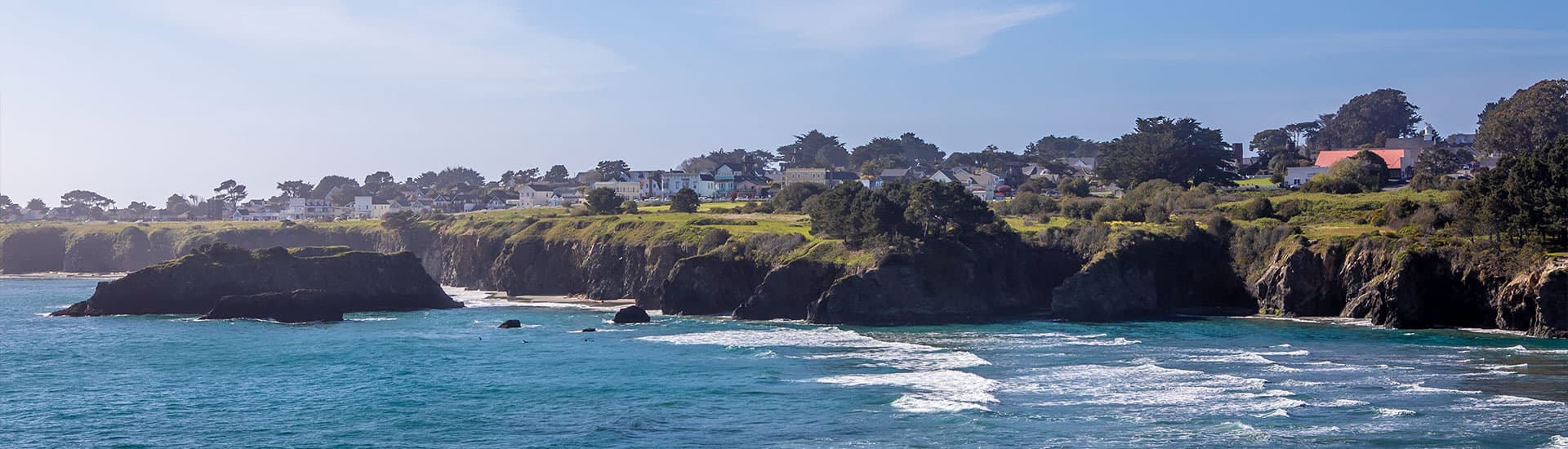 Coastal landscape with cliffs, ocean waves, and a small town in the background under a blue sky.