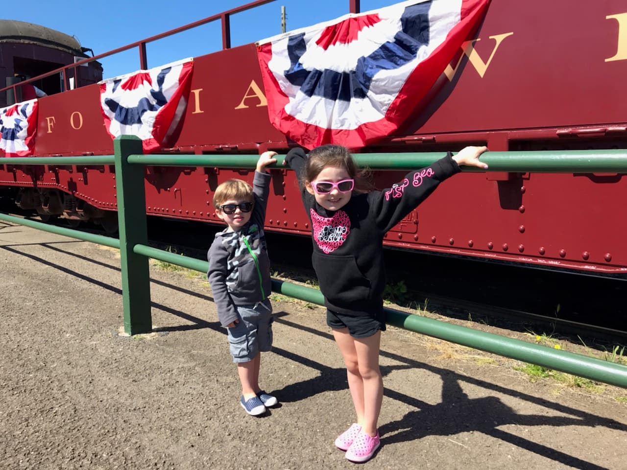 Two kids in sunglasses pose by a vintage train decorated with red, white, and blue bunting. Two kids in sunglasses pose by a vintage train decorated with red, white, and blue bunting.