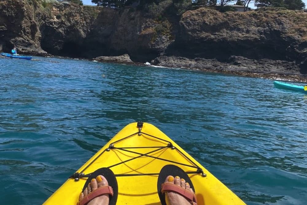 Feet resting on a yellow kayak with a view of rocky cliffs and other kayakers in the background.