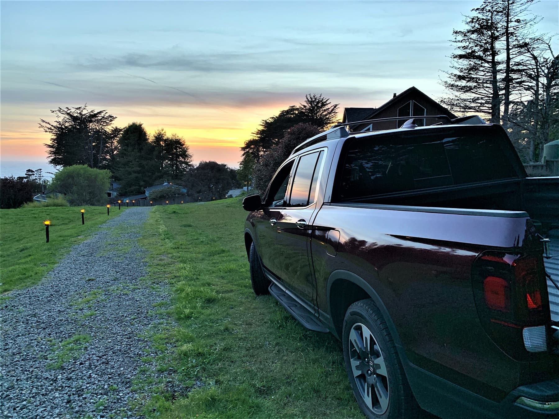 A dark red pickup truck parked on a gravel path with a sunset backdrop and trees in the distance.