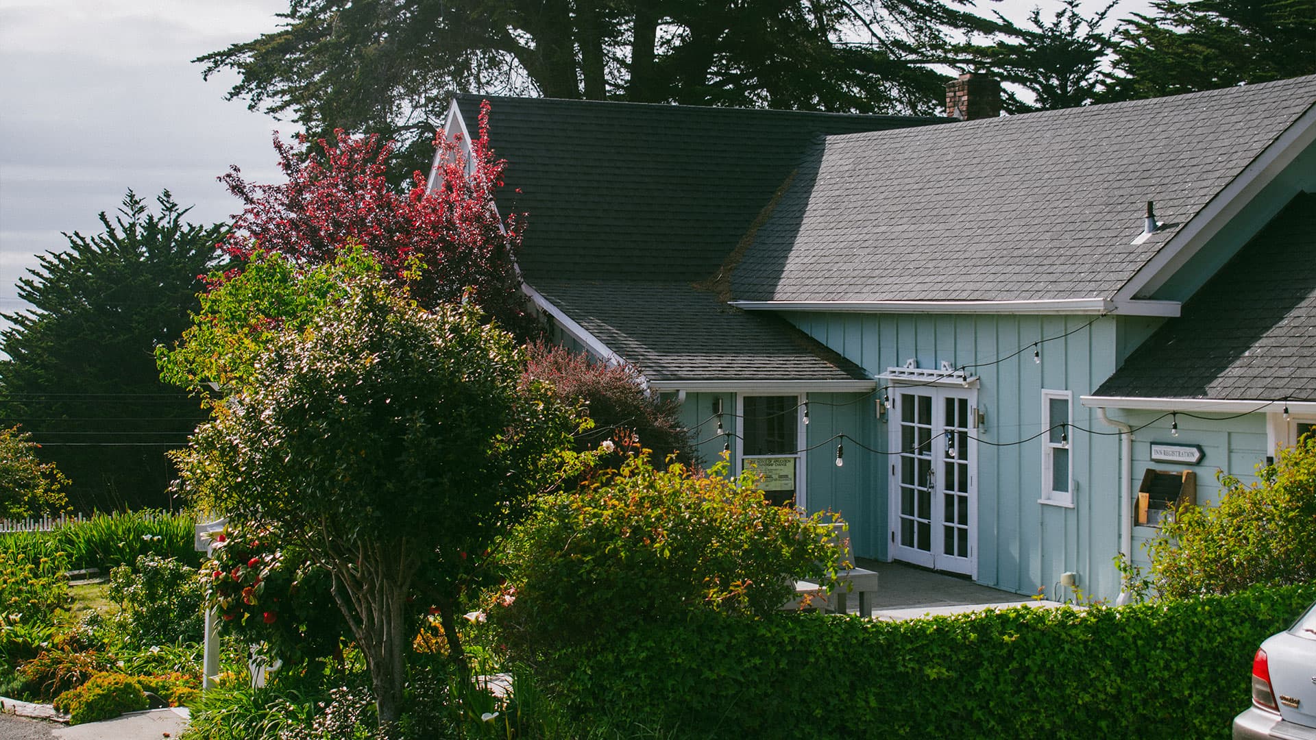 Exterior of a charming light blue cottage with lush greenery, trees, and flowering bushes surrounding the entrance.