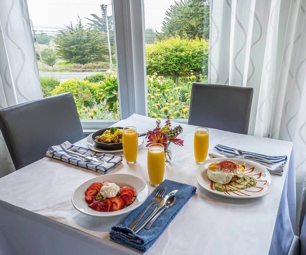 A beautifully set breakfast table with two plates of food, three glasses of orange juice, and a view of a garden through a large window.