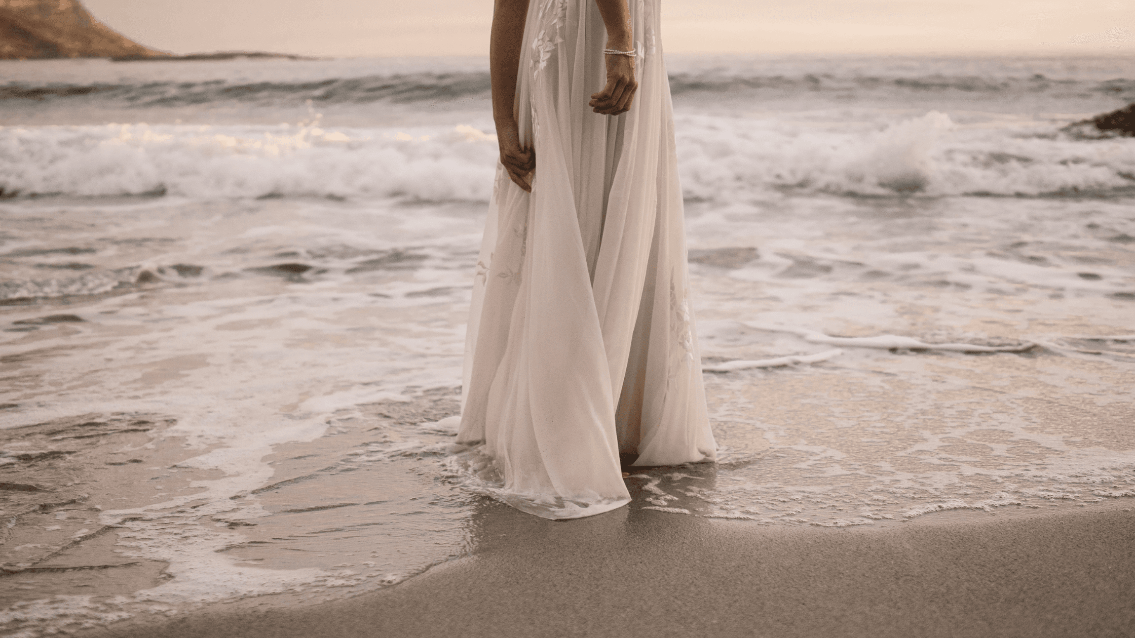 A woman in a flowing white dress stands at the water's edge, waves gently lapping at her feet.
