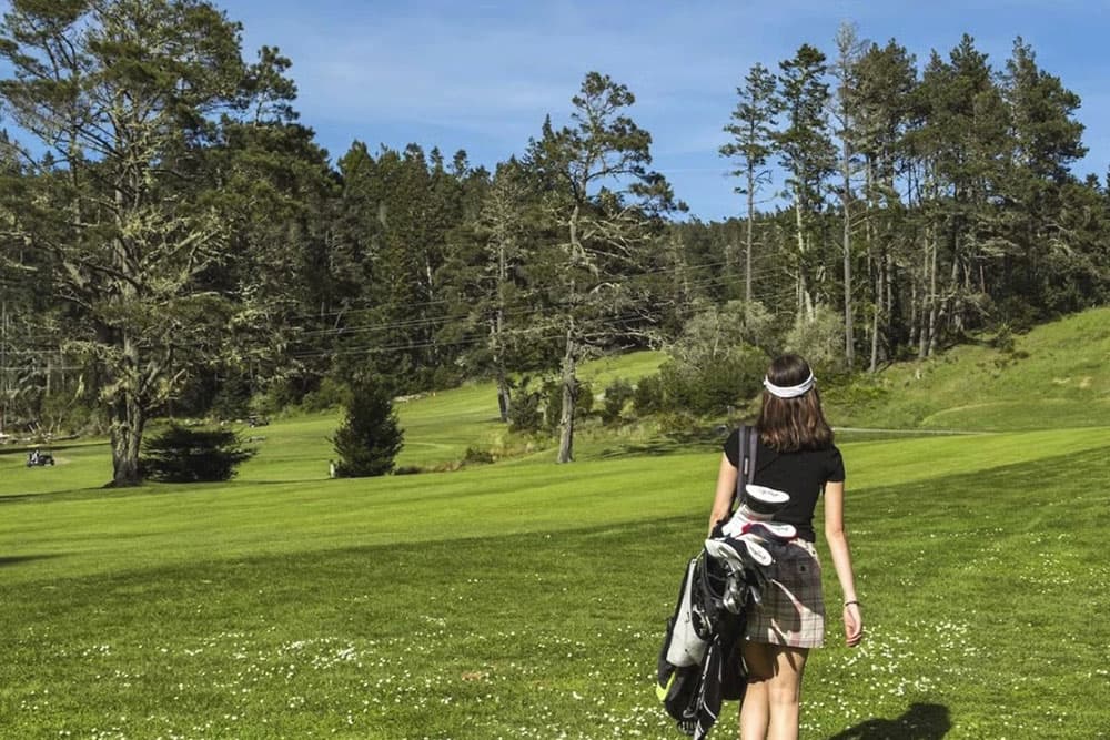 A golfer walks toward a lush green course, carrying clubs and wearing a visor.