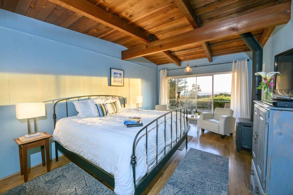 A cozy bedroom featuring a metal bed frame, wooden beams, and large windows that let in natural light.