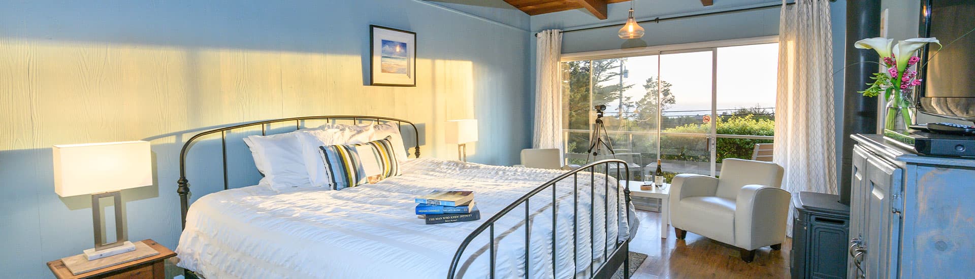 A cozy bedroom featuring a metal bed frame, bedside lamps, and large windows with a view of greenery.