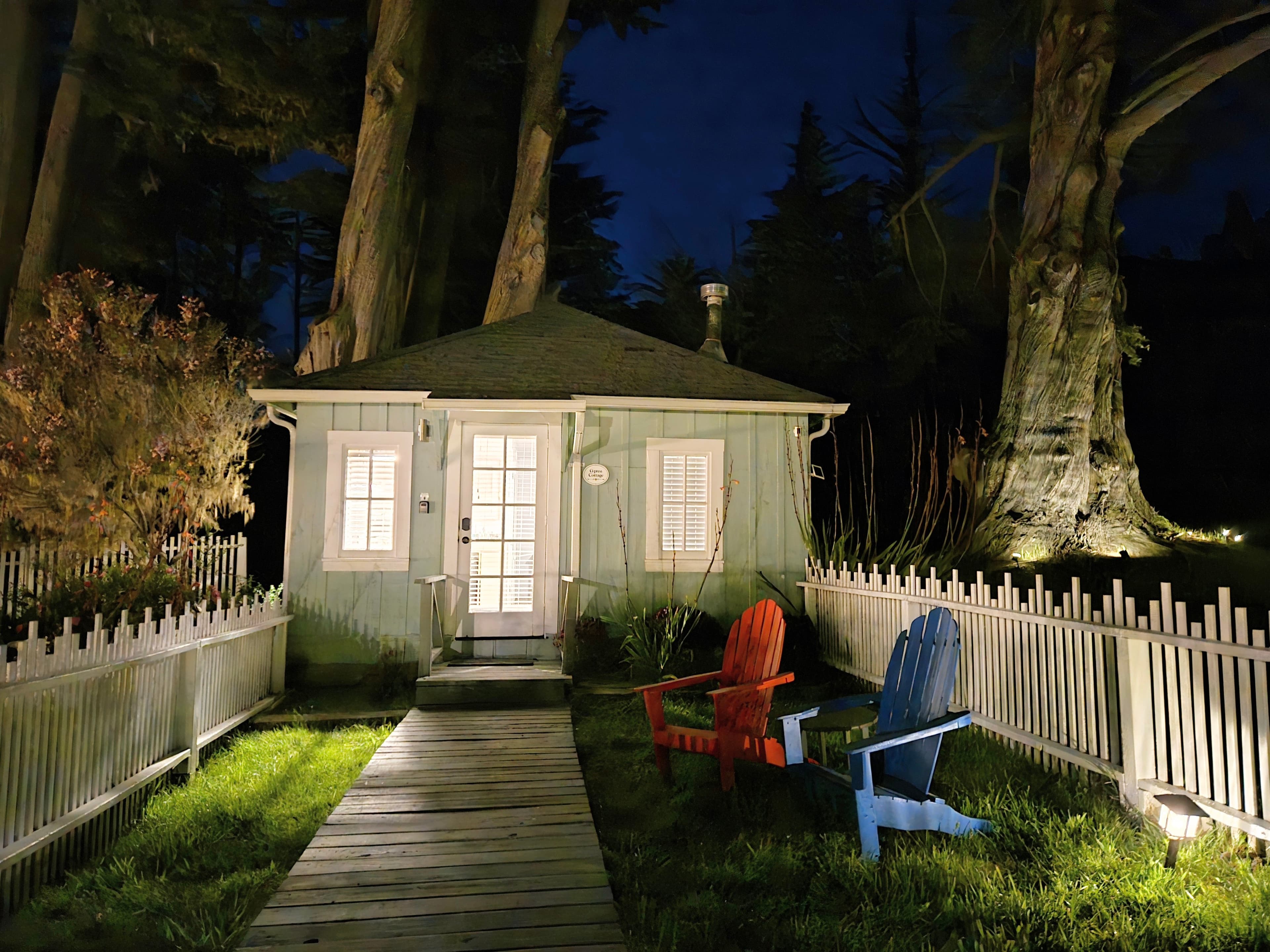A cozy, lit cabin at night surrounded by trees, with a wooden pathway leading to it and colorful adirondack chairs in the foreground.
