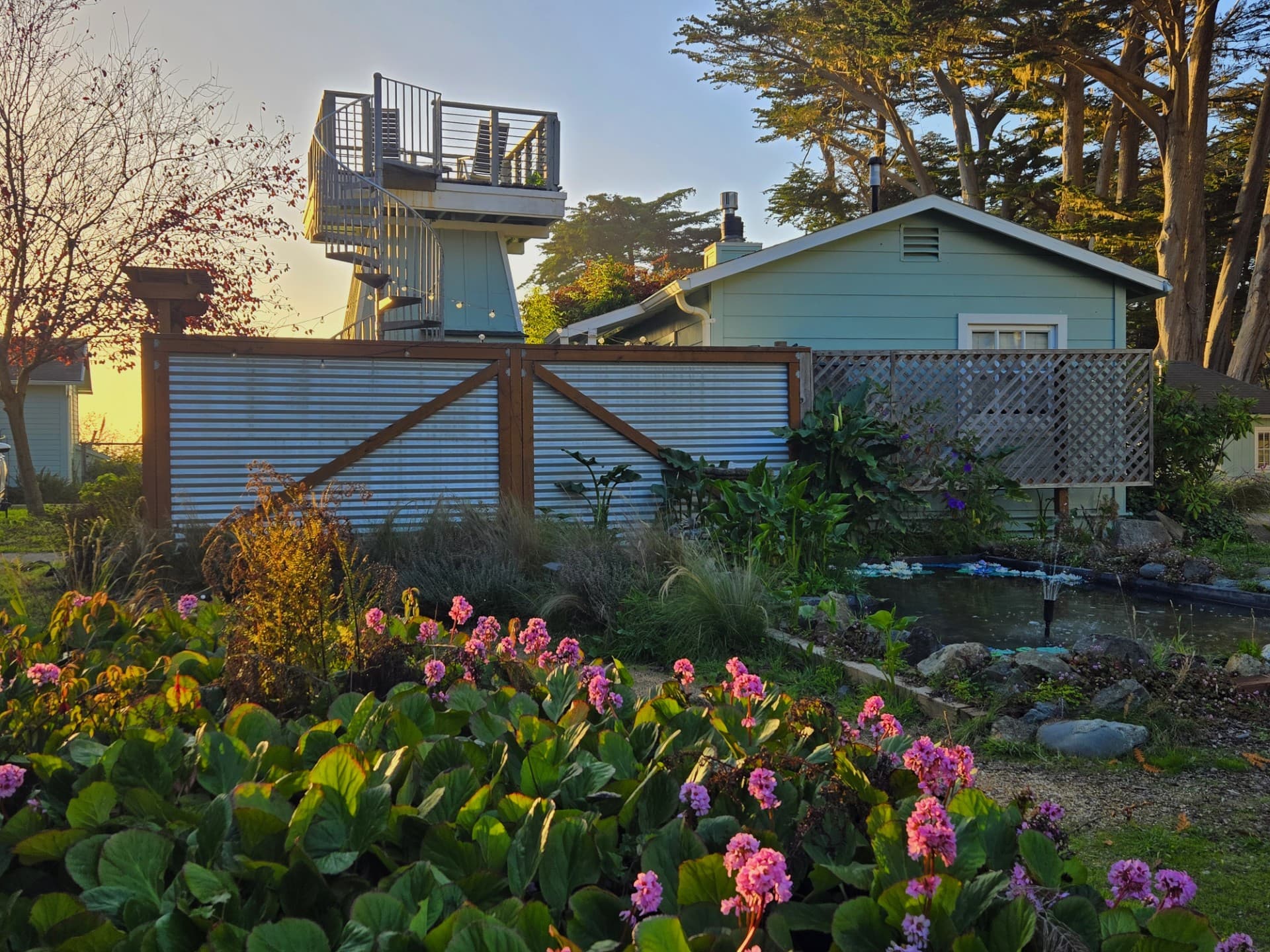 A tranquil garden with vibrant flowers in the foreground, featuring a blue house and a lookout tower in the background under a clear sky.
